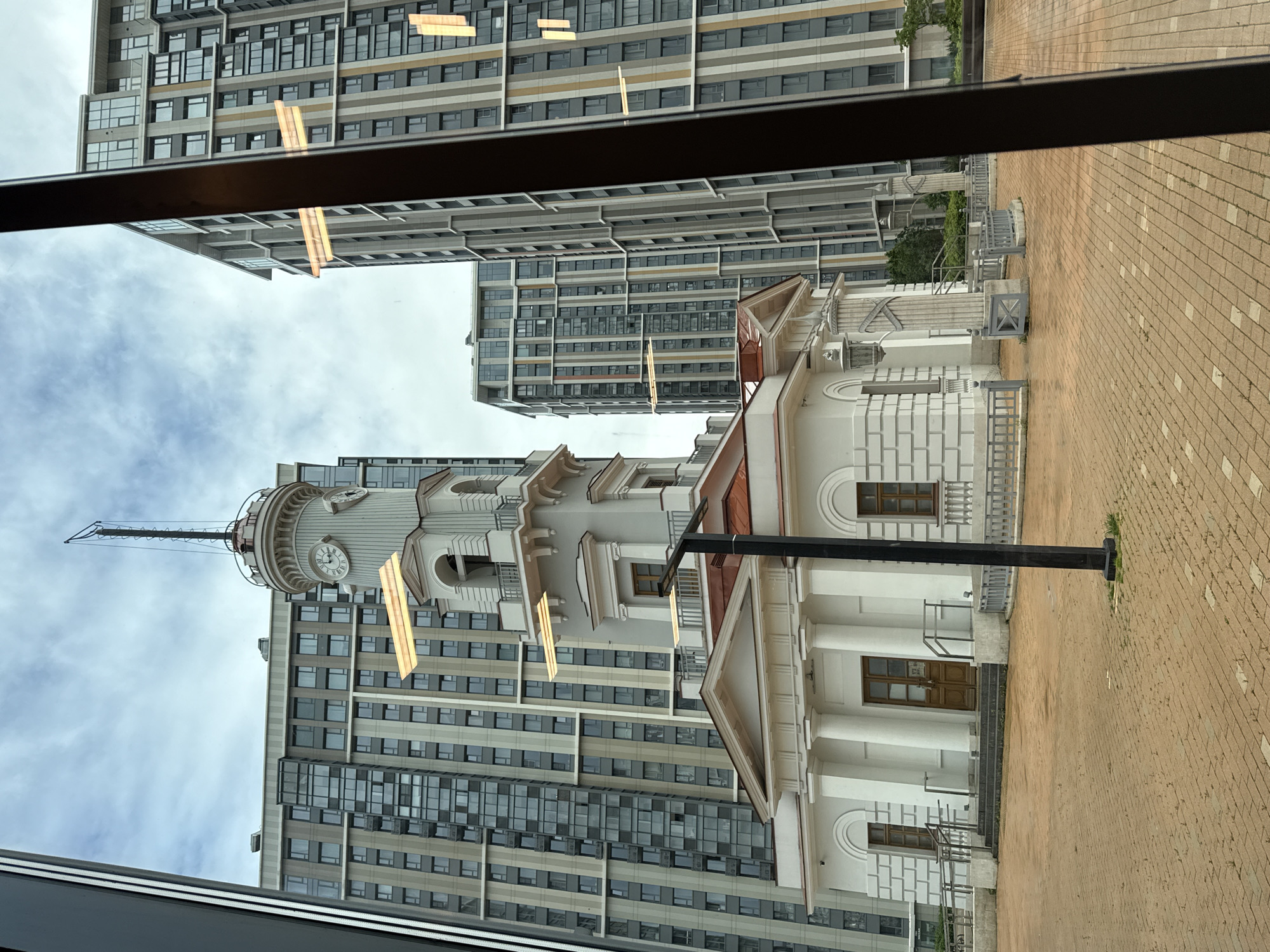 The image depicts the Church of the Holy Martyr Tatiana of Rome at Moscow State University, Moscow, Russia, as seen from inside a building through a window. The church is a white building with a tower and a clock on its facade. It is located on a brick-paved ground. Behind the church are modern residential apartment buildings with numerous windows. The sky is overcast.