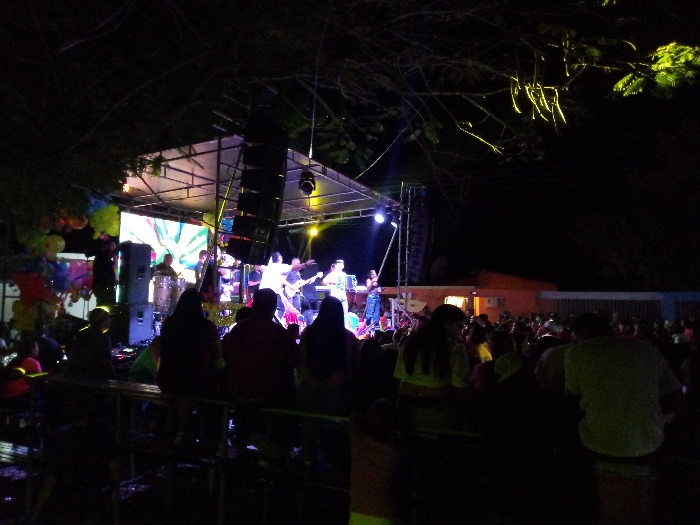 A band performs on an outdoor stage at night. The stage is lit with bright lights and has a large screen behind the performers. A crowd of people sits on benches and stands watching the show. The sky is dark, and there are trees in the background. An orange building is visible to the right.
