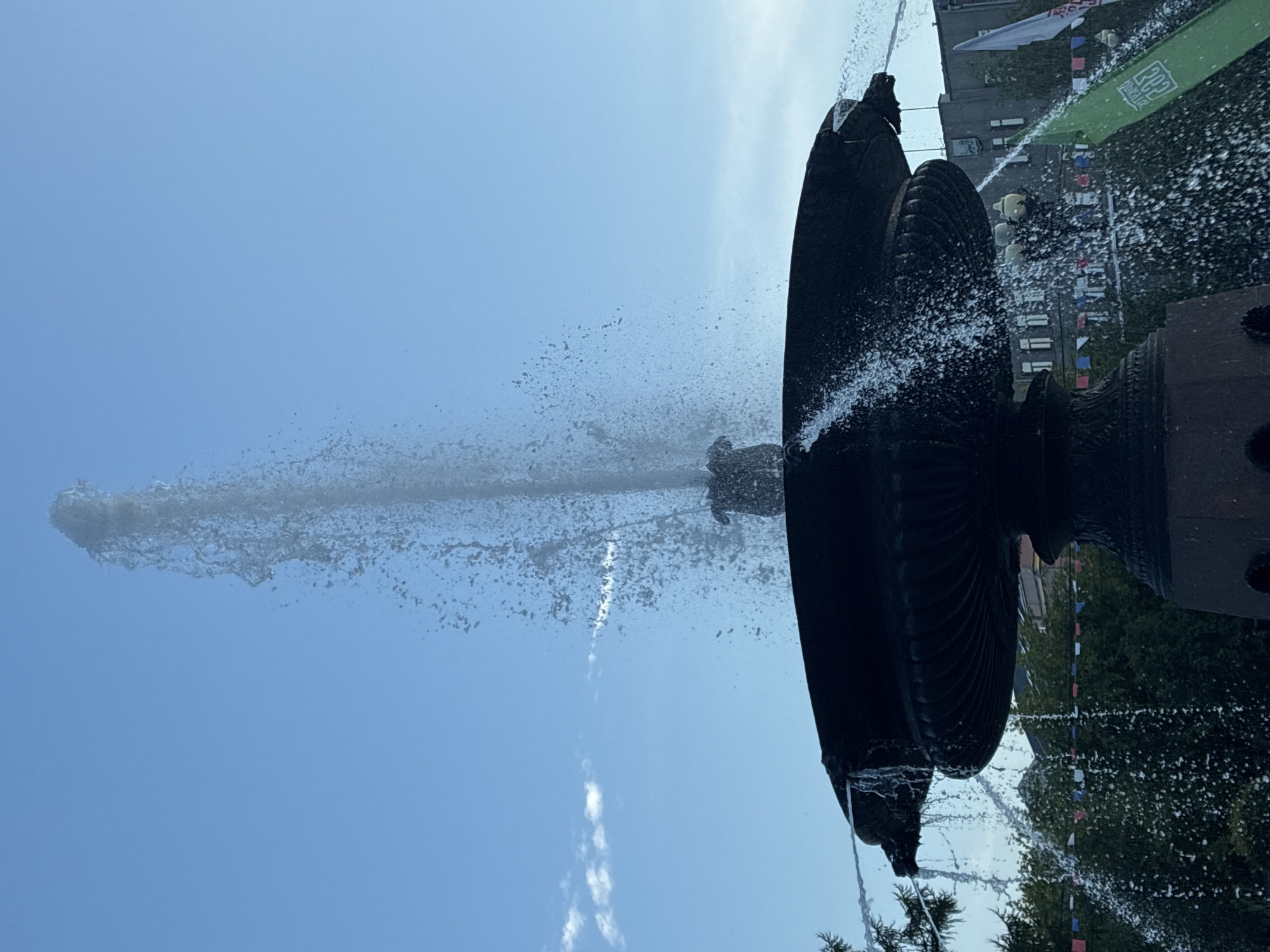 The image shows a close-up view of a large, dark-colored stone fountain spraying water against a bright blue sky with a few wispy clouds. The fountain has multiple tiers, with water cascading down the sides and a powerful jet shooting upwards from the center. In the background, there are trees and buildings visible, suggesting an urban setting. String lights with small flags are strung between trees, and a white flag with text is visible on the right side of the frame. The overall scene captures a moment of dynamic water movement in an outdoor public space. This fountain is located in the Kirov Square (Ploshchad Kirova) in Irkutsk, Russia.