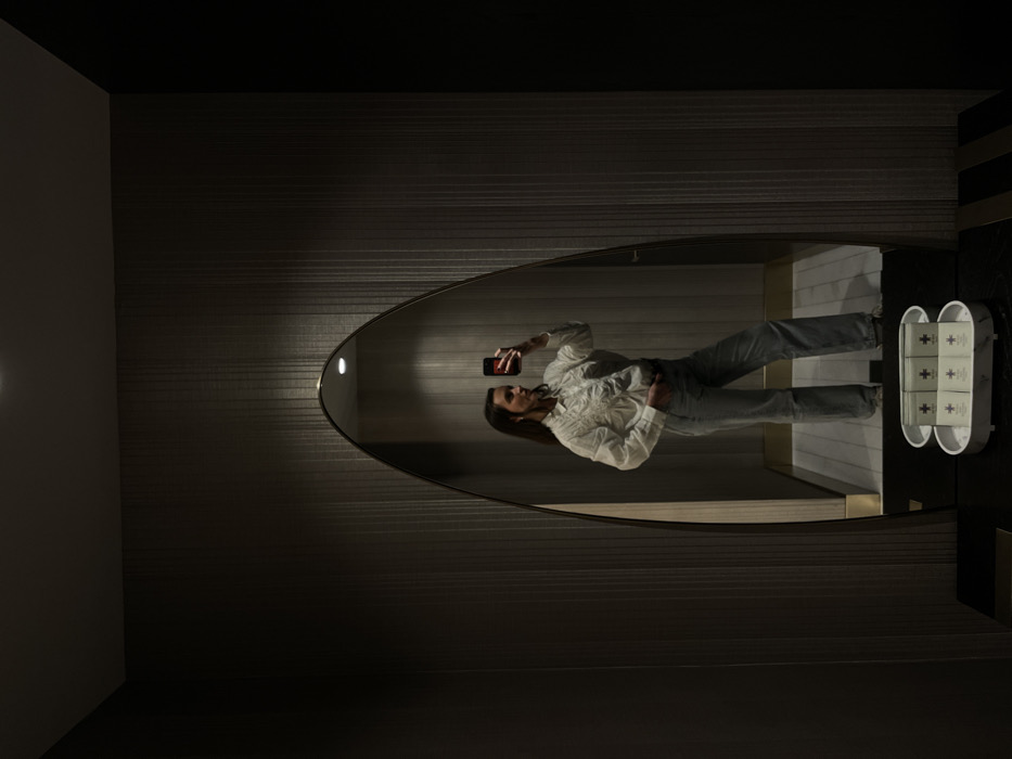 A woman is reflected in a curved mirror in a dimly lit room with horizontal striped wallpaper. She's holding a phone, presumably taking a picture. She wears a white shirt and jeans. A white dispenser with three compartments, each marked with a cross, sits on a black surface to the right of the mirror. The room has dark corners.
