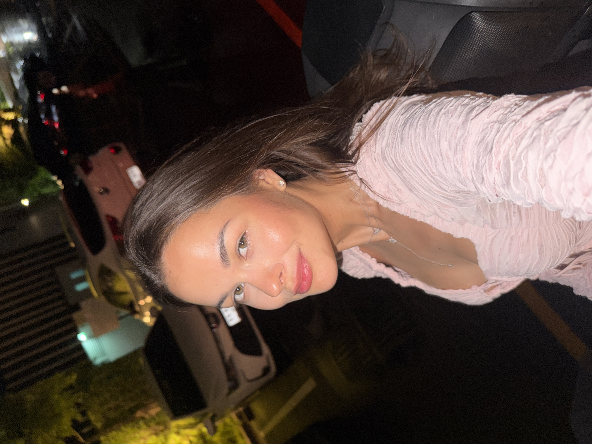 A young woman with long, dark brown hair and light-colored eyes is captured in a selfie, looking directly at the camera with a subtle smile. She is wearing a light pink, textured, off-the-shoulder top. Her facial features are clear, with natural-looking makeup, slightly rosy cheeks, and full, light pink lips. A delicate silver chain necklace is visible around her neck, and a small stud earring adorns her ear.
The image appears to be taken at night, indicated by the dark background visible through what seems to be a vehicle window. Blurred lights, reflections, and indistinct shapes of buildings or other vehicles are visible in the background, suggesting an urban or suburban setting. A light-colored object, possibly a cell phone, is partially visible in the lower left background, reflecting some light. Parts of the vehicle's interior, such as a dark seat or dashboard, are also visible, framing the woman. The overall impression is that she is posing inside a car or similar vehicle at night, perhaps in Choeng Thale, Thailand. No visible text is present in the image.