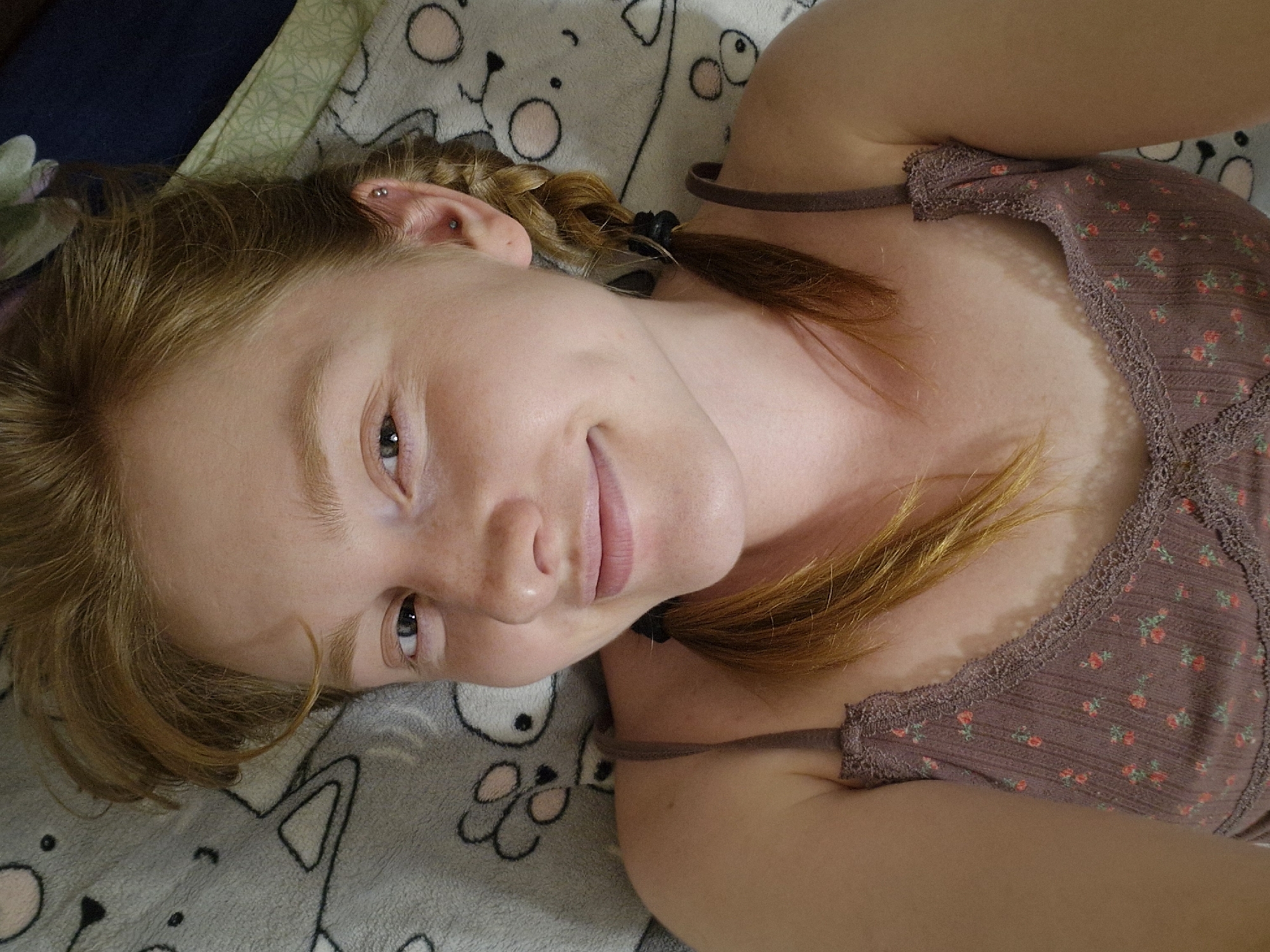 A young, light-skinned person with strawberry blonde hair in pigtails lies on a gray blanket patterned with cartoon animal faces. They are wearing a brown floral-patterned top with thin straps. The person is looking up at the camera with a slight smile. The setting appears to be indoors. No specific location in Yekaterinburg, Russia, can be determined from the image.