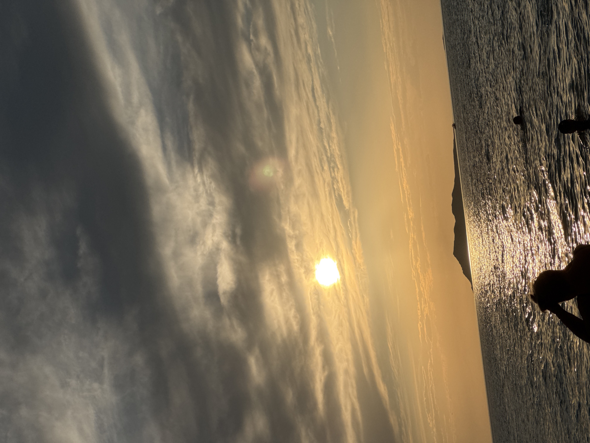 This is a photograph of the Caspian Sea at sunset, near the village of Zharbulak, Kazakhstan. The sun is low in the sky, casting a warm, golden glow on the water. Clouds are scattered across the sky, with some darker clouds in the upper left corner and lighter clouds illuminated by the sun. In the distance, a silhouette of a landmass, likely an island or peninsula, is visible. The water is choppy, reflecting the sunlight. In the foreground, the silhouette of a person's head and shoulders is visible, with their hand raised to their head. Further out in the water, there are at least two other people swimming. The overall scene depicts a tranquil, natural setting during twilight.