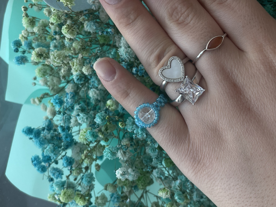 A close-up photograph depicts a human hand, specifically the dorsal side, holding a bouquet of flowers. The hand, featuring light skin tone, has four fingers visible. The index finger displays a ring crafted from small blue beads surrounding a clear, circular stone or bead. The middle finger wears two silver-toned rings; the upper ring features a heart-shaped white inlay bordered by small clear stones, while the lower ring has a large, rectangular, clear faceted stone. The ring finger has a thin, silver-toned band with an elongated oval, reddish-orange stone. The background consists of a dense arrangement of small, clustered flowers, primarily light blue and white or pale green, consistent with dyed baby's breath (Gypsophila). The flowers are partially wrapped in a teal-colored paper or cellophane visible in the upper left. The image provides no discernible visual information to indicate the specified city location of Соболиха, Russia.