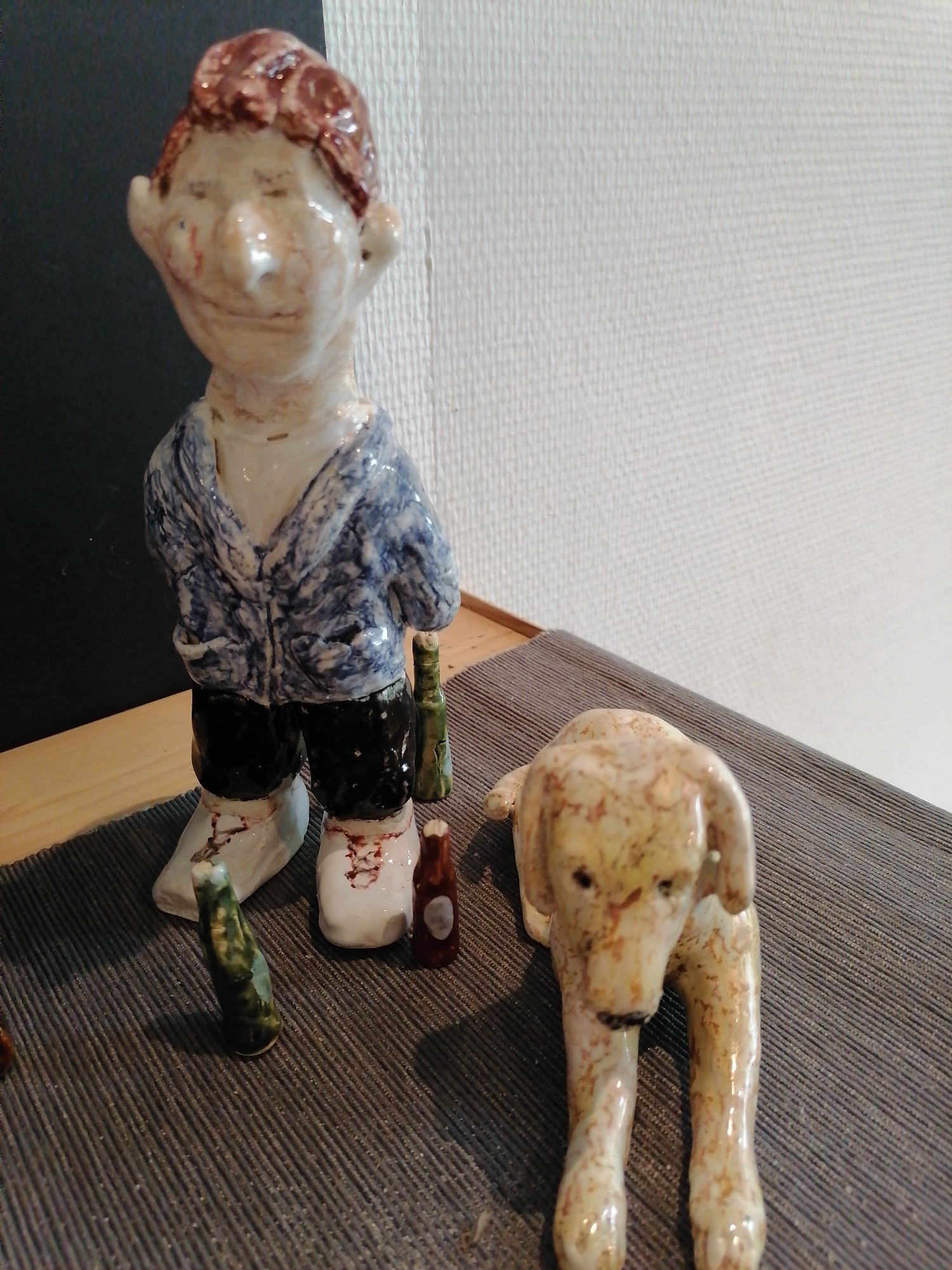 This is a close-up, eye-level, indoor shot of a collection of ceramic figurines. In the center-left, a roughly formed ceramic figurine of a person stands upright. The figurine has a pale skin tone with reddish-brown hair and appears to be wearing a blue and white patterned jacket over black pants and light-colored shoes. The figurine is holding a green bottle in its right hand. To the right of the standing figurine, a pale yellow ceramic dog figurine lies down. The dog has brown speckles and appears to be looking forward. In the foreground, between the two main figurines, are two small ceramic bottles, one green and one reddish-brown. The figurines are placed on a dark grey ribbed mat. Behind the figurines is a wooden surface, and further back, a white textured wall. The scene is lit with artificial light. The location is not specified in the image.