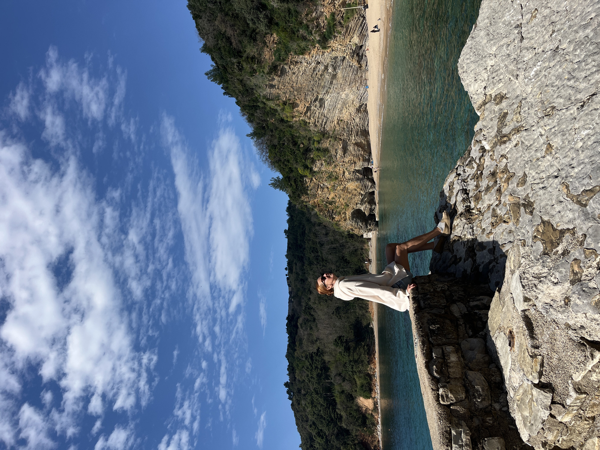 A scenic shot depicts a woman sitting on a rocky ledge overlooking the Adriatic Sea. The scene appears to be in the city of Budva, Montenegro, evidenced by the coastal landscape. The woman wears a light-colored top and sunglasses, facing towards the sea with a relaxed posture. The background includes a beach, cliffs covered in vegetation, and a blue sky with scattered clouds. The image has a slight tilt, with the horizon line appearing diagonal. The sea is a vibrant blue-green color, contrasting with the rocky ledge in the foreground.