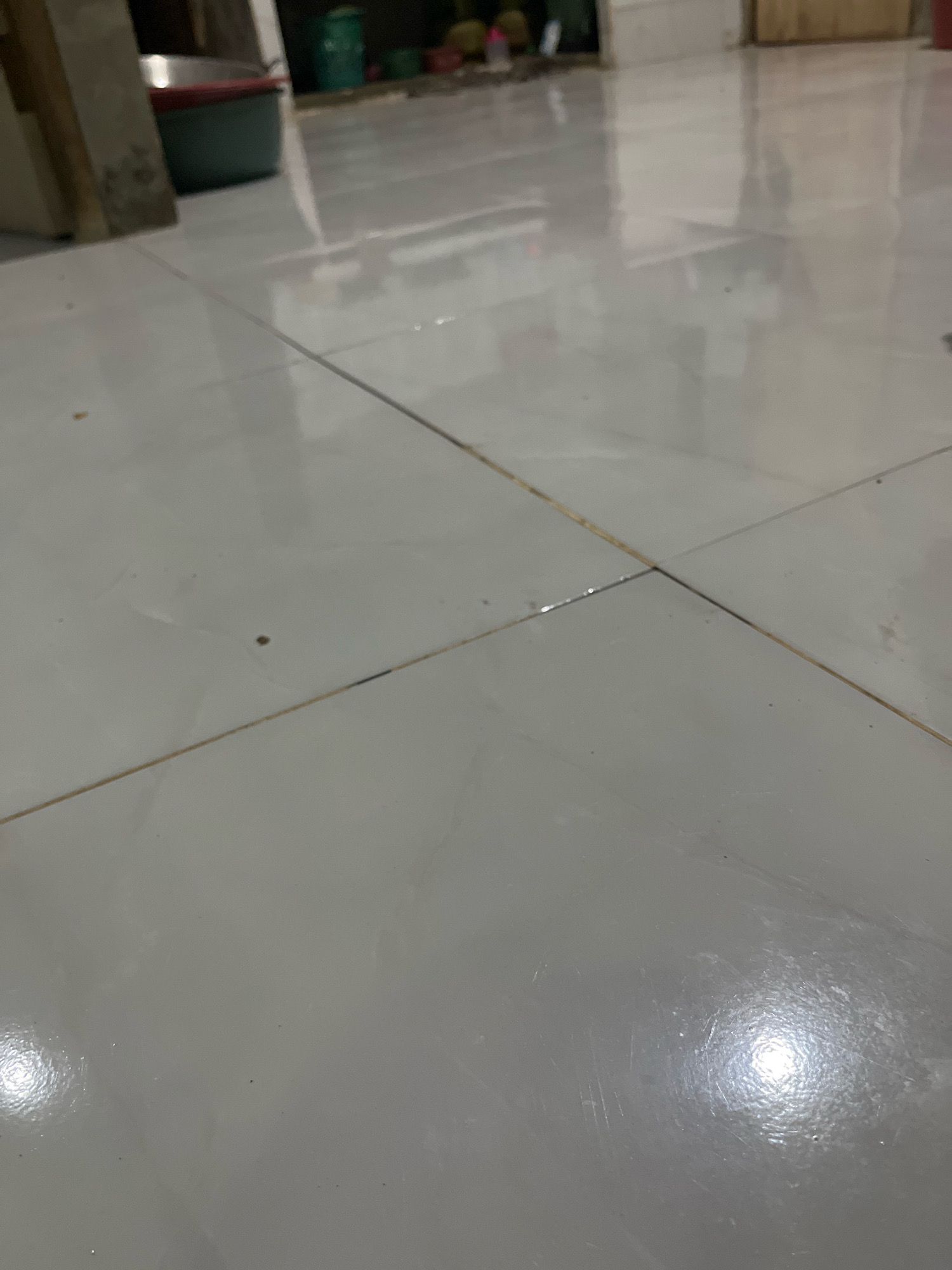 The image depicts a tiled floor with visible grout lines. In the background, a blurry scene shows what appears to be a storage area with containers, possibly in a residential or commercial setting. The lighting suggests an indoor environment with overhead illumination creating reflections on the glossy tiles. The overall impression is one of a functional, everyday space.
