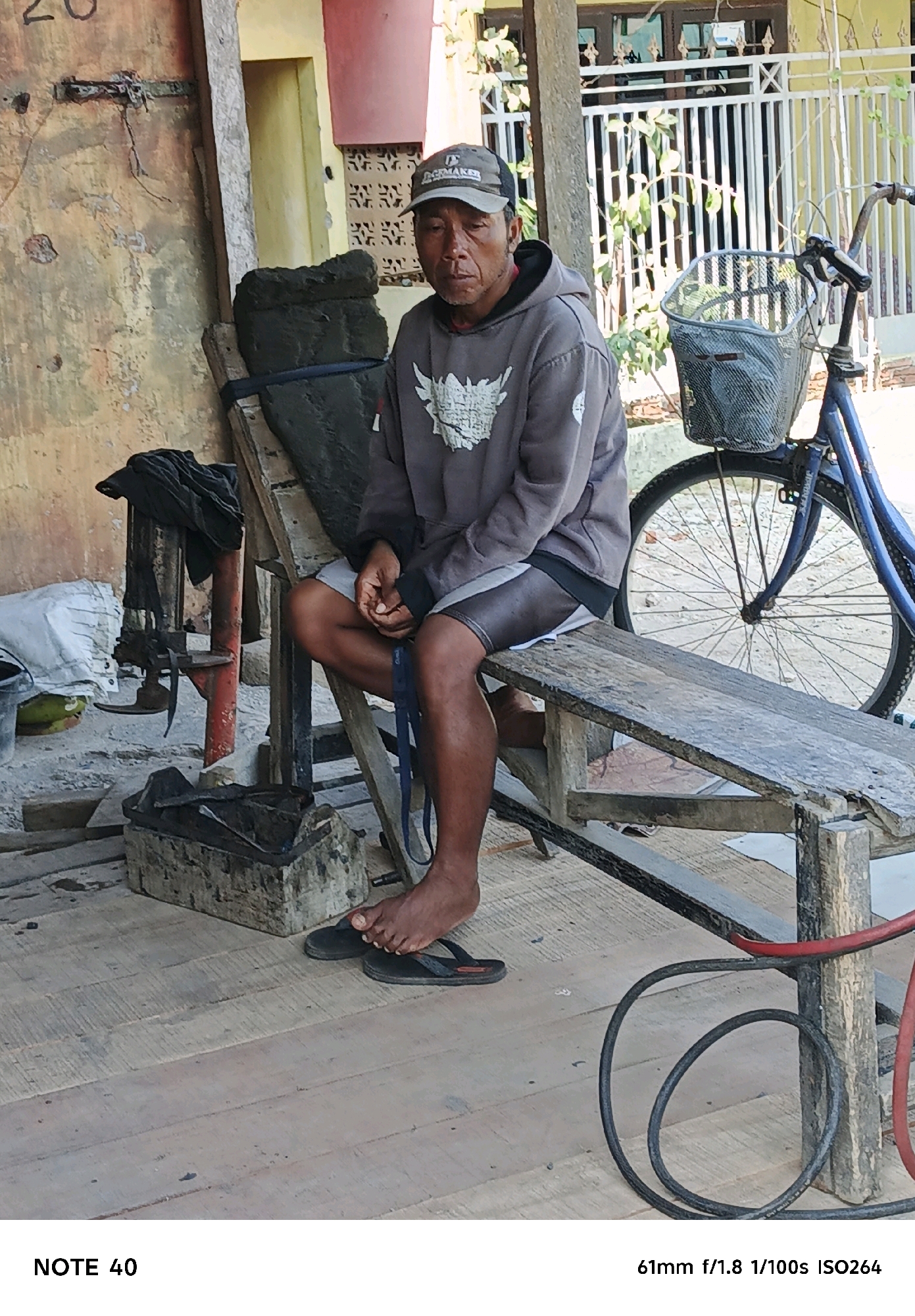 An older Indonesian man wearing a grey hooded sweatshirt and striped shorts sits on a wooden bench in an outdoor setting. He has a baseball cap on and appears to be looking down. A bicycle with a basket is positioned to his right. The background shows a building with yellow and pink walls, a white metal fence, and vegetation. The foreground consists of a wooden plank floor and various objects including a piece of black material, a metal device, and coiled black hoses. The scene suggests a casual or perhaps work-related environment in a local community.