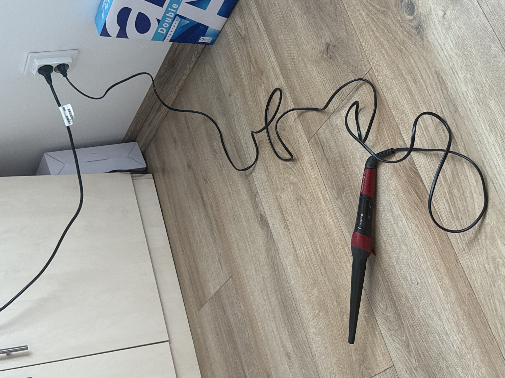The image shows a curling iron plugged into a wall socket. The curling iron is black with a red handle and lies on a wood-patterned surface. The curling iron's cord is black and tangled. A white box and a box with text visible ("Double A...") are situated near the wall socket. The wall socket is white and recessed into a white ceiling. A white cabinet with a metal handle is visible in the lower left of the image. The setting appears to be indoors. Given the presence of a European-style plug, the image is plausibly located in Sofia, Bulgaria, but this cannot be definitively determined from the image alone.