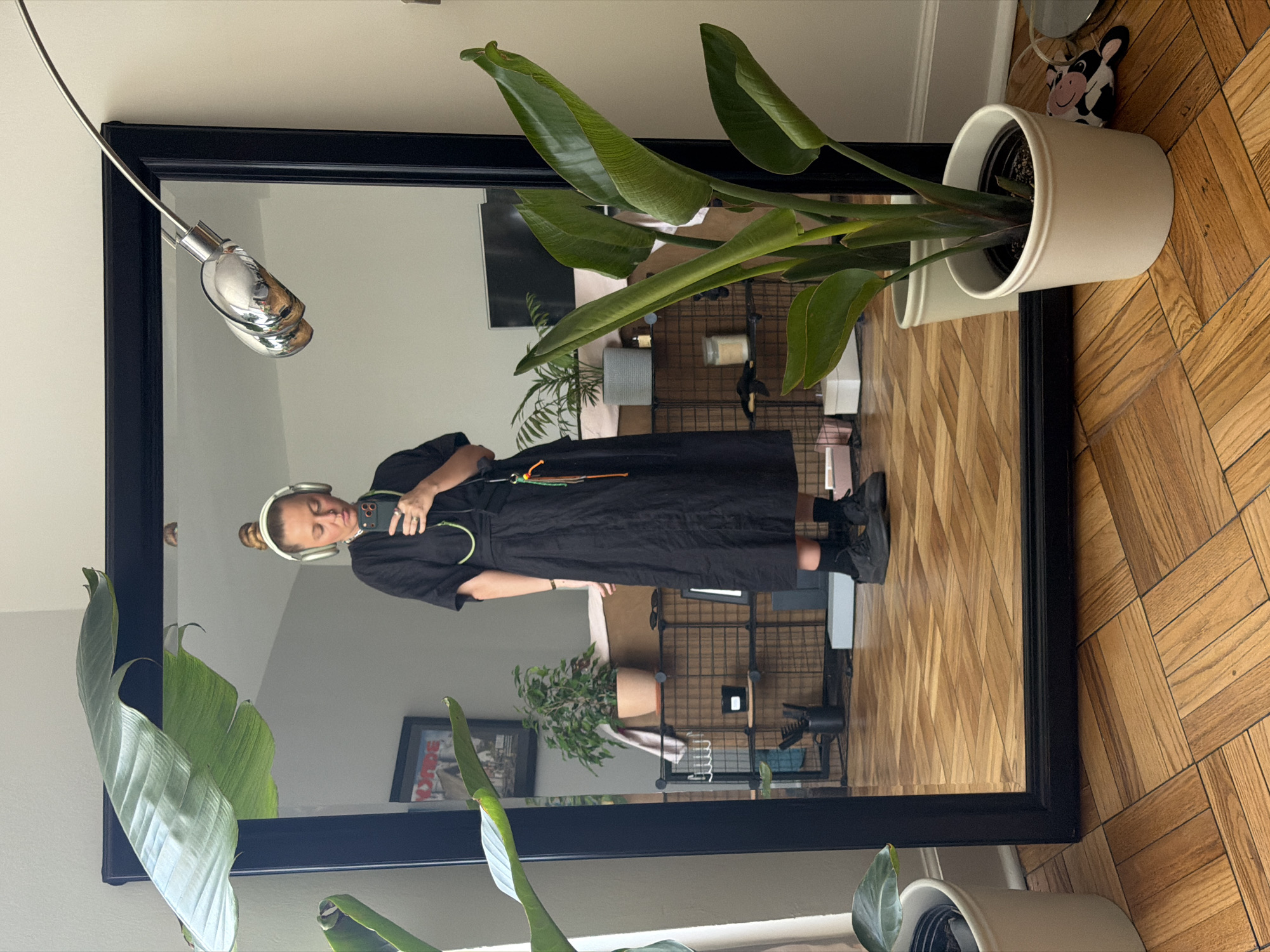 A person wearing headphones and a black dress is reflected in a large, black-framed mirror. The person is holding a smartphone, seemingly taking a photo. The mirror's reflection reveals a section of a room, including shelving with various items. Outside the mirror's frame, a large Bird of Paradise plant sits in a white pot, both at the top and bottom of the frame. The floor is herringbone-patterned wood. The city of Los Angeles, United States of America, is not explicitly discernible from the visual information provided.