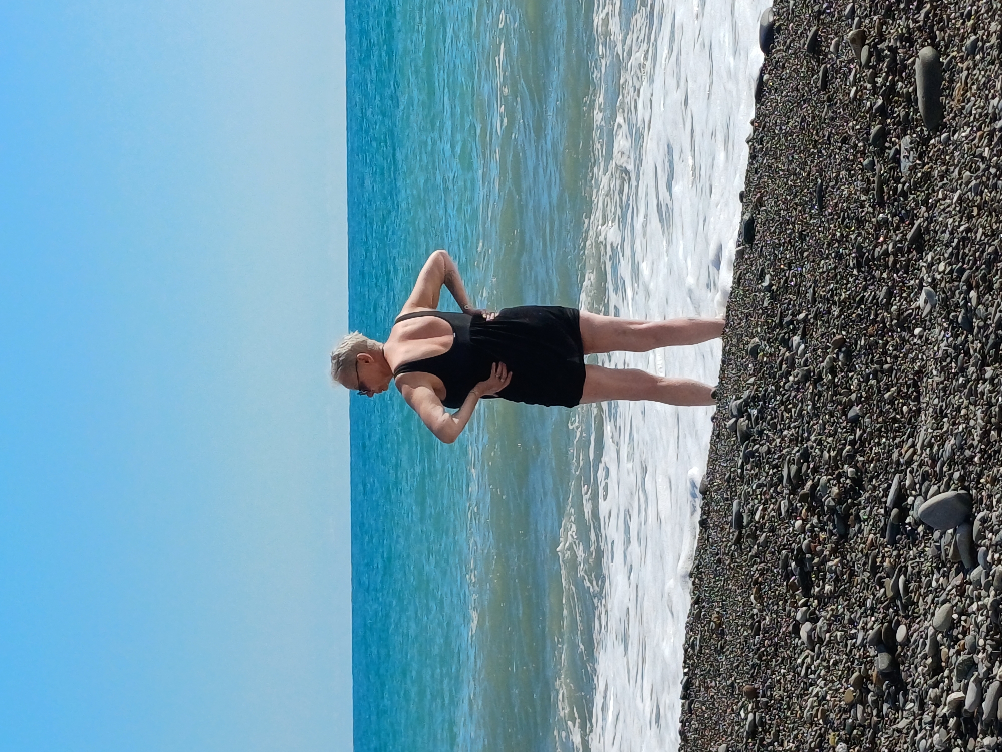 A middle-aged woman with short, grey hair and sunglasses stands on a pebble beach, facing away from the camera and towards the sea. She is wearing a black one-piece swimsuit. Her hands are on her hips, and she appears to be looking out at the water. The sea is a vibrant blue-green, with gentle waves lapping at the shore. The sky above is clear and blue. The pebble beach is covered in various shades of grey and brown stones. The scene is likely located in Sochi, Russia, given the clear blue sky and the characteristic pebble beaches found in the region.
