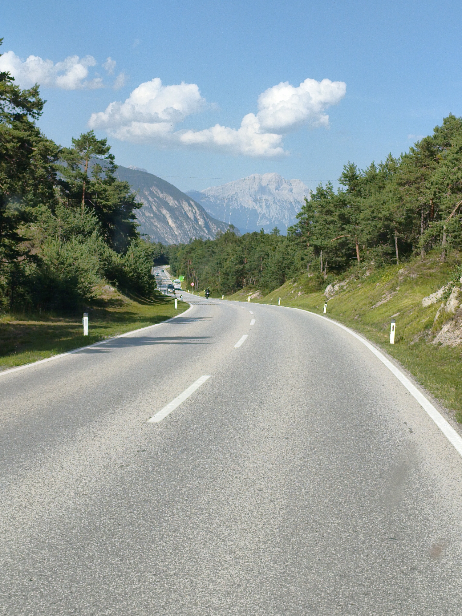 The image shows a winding asphalt road in a mountainous, forested area. The road has white lane markings and is bordered by grassy shoulders with some rocks and white marker posts. On the left side of the road, there are dense evergreen trees, and on the right side, the terrain slopes upwards, also covered in trees and grass. In the distance, the road continues to curve, with a few vehicles visible, including a green bus and what appears to be a motorcycle. Beyond the trees, a range of mountains with rocky peaks and snow in some areas is visible under a bright blue sky with scattered white clouds. This scene is consistent with a road in the vicinity of Sautens, Austria, given the characteristic Alpine landscape.