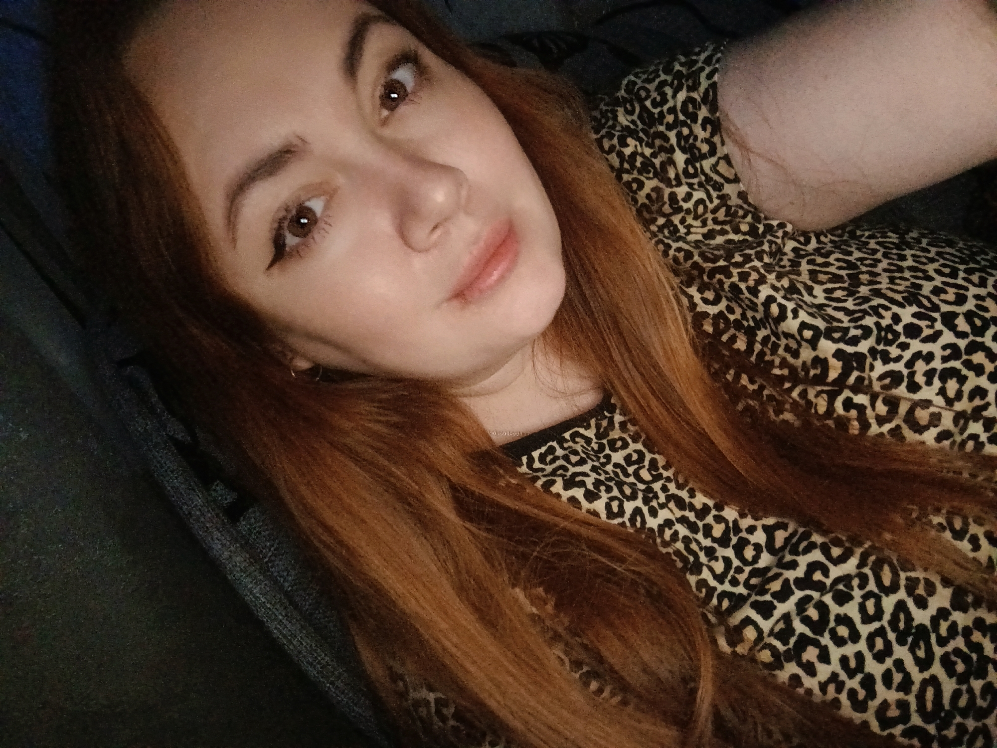 A close-up, softly lit indoor portrait features a young woman with long, flowing reddish-brown hair. She has brown eyes, accentuated by winged eyeliner, and a subtle, relaxed expression on her face, with slightly parted lips. She appears to be reclining or lying down, looking directly into the camera. She is wearing a black and beige leopard print top. Her hair cascades around her shoulders and across her chest, intermingling with the pattern of her clothing. The background consists of dark, soft fabric, likely a couch or bedding, contributing to the intimate and cozy feel of the scene. The lighting is dim, suggesting it might be evening or night. There are no specific location cues visible in the image to confirm the provided context of Chaikovsky, Russia, nor is any text visible.