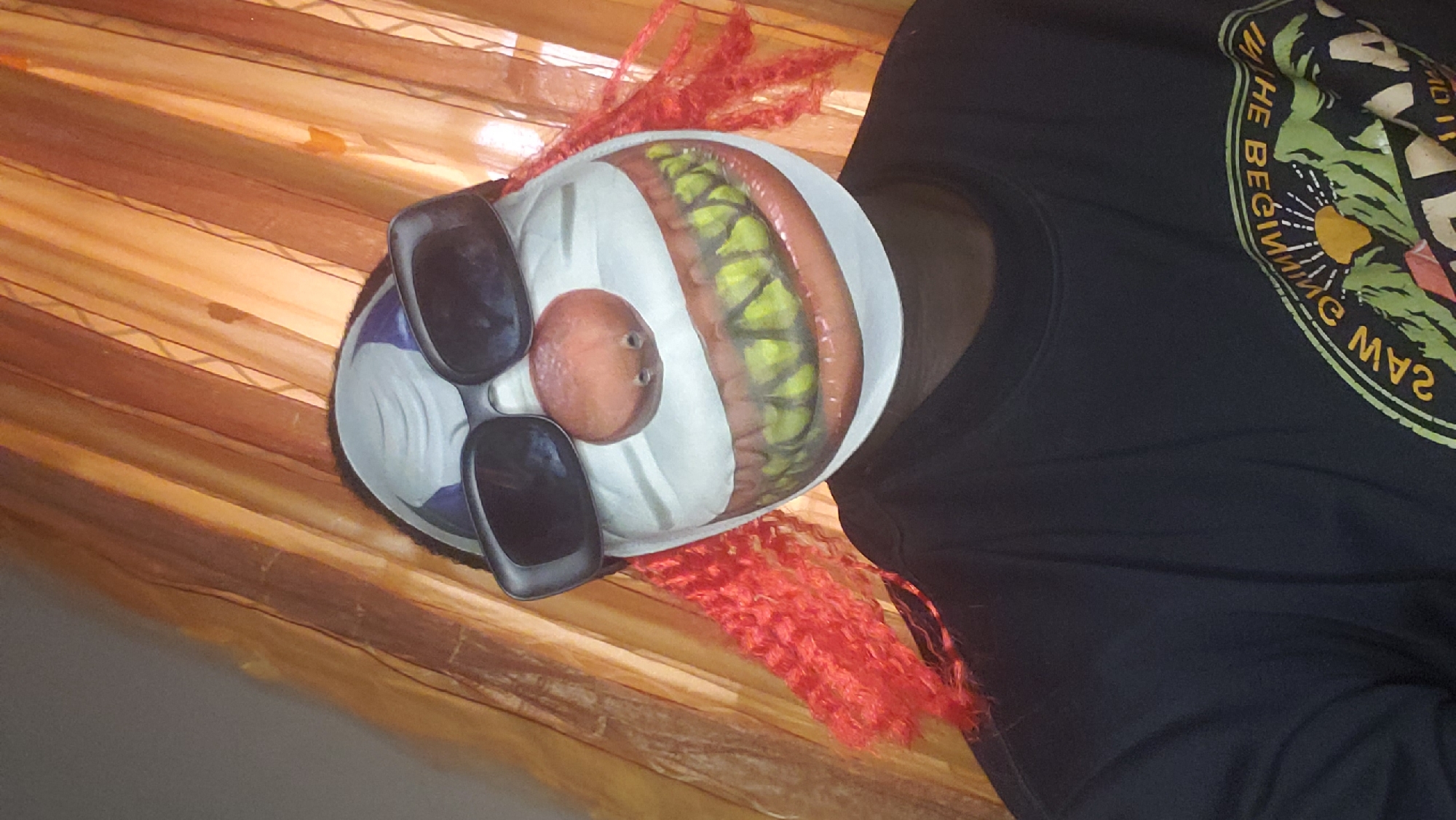 A clown mask with black sunglasses and orange hair sits on a striped surface next to a person wearing a black shirt with a graphic design. The location is not identified in the image, but the user asked for Sabon Tasha, Nigeria.