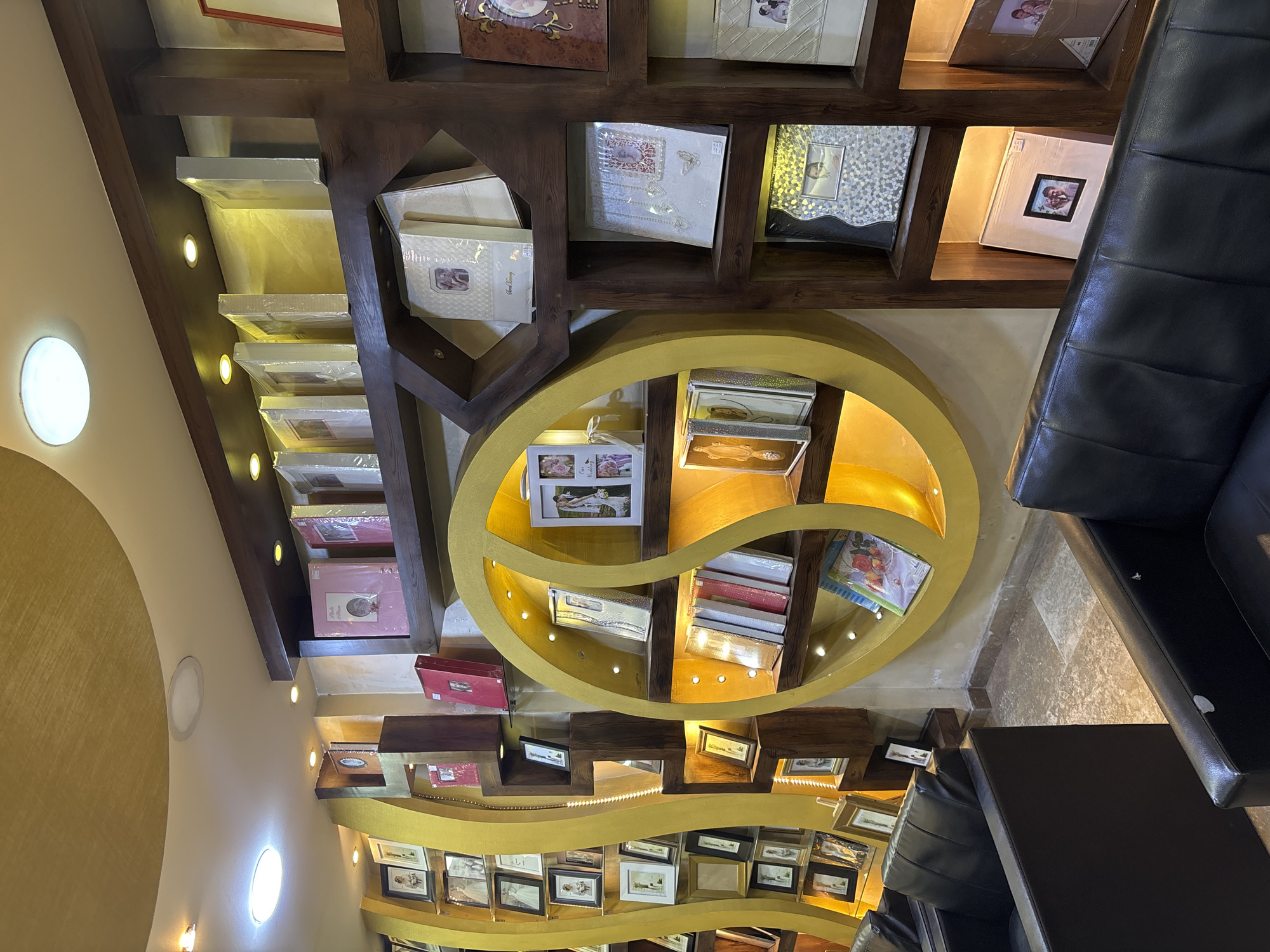 The image depicts the interior of a retail establishment, likely a photo album or framing shop, located in Amman, Jordan. The store features extensive shelving units displaying a variety of photo albums, picture frames, and keepsake boxes. The decor includes warm, golden-toned lighting and wooden accents, creating an inviting atmosphere.
In the foreground, two black leather sofas are visible, suggesting a waiting area or customer lounge. The shelves are organized with albums of different sizes and colors, some appearing to be new and still in protective plastic wrapping. There are also individual picture frames, some empty and others containing photographs.
A prominent circular shelving unit, finished in a bright yellow, is a focal point of the display. This unit has multiple tiers holding more albums and frames. The overall impression is one of a well-stocked store specializing in photographic mementos. No people are visible in the image.