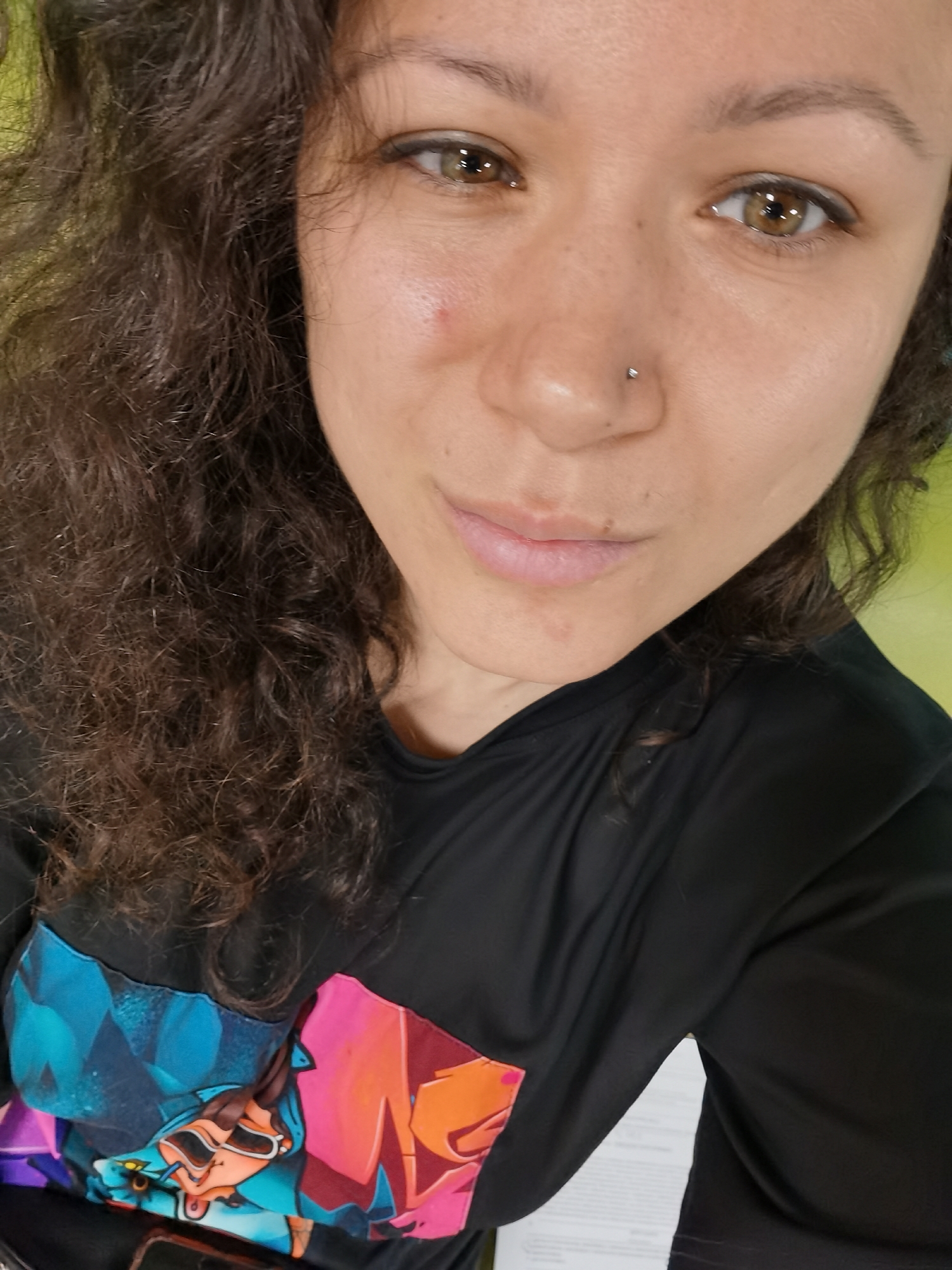 The image presents a medium close-up, self-portrait of an adult female. She has a fair to medium complexion, dark brown curly hair that falls over her left shoulder, and brown eyes with visible amber flecks. Her upper eyelids are lined with dark eyeliner. A small silver stud is present in her left nostril piercing. A subtle reddish blemish is visible on her right cheek, and several small moles or freckles are present on her face. Her lips are light pink and slightly parted, contributing to a neutral or mildly contemplative expression. She is wearing a long-sleeved black top featuring a vibrant, multi-colored graphic print across the chest, composed of abstract or stylized elements in shades of blue, orange, pink, and purple. The background is significantly out of focus, showing a soft green blur on the left side and a lighter, indistinct blur on the right. Portions of a white paper document or material are visible in the lower right foreground, below her shirt. The scene is situated in Izhevsk, Russia.