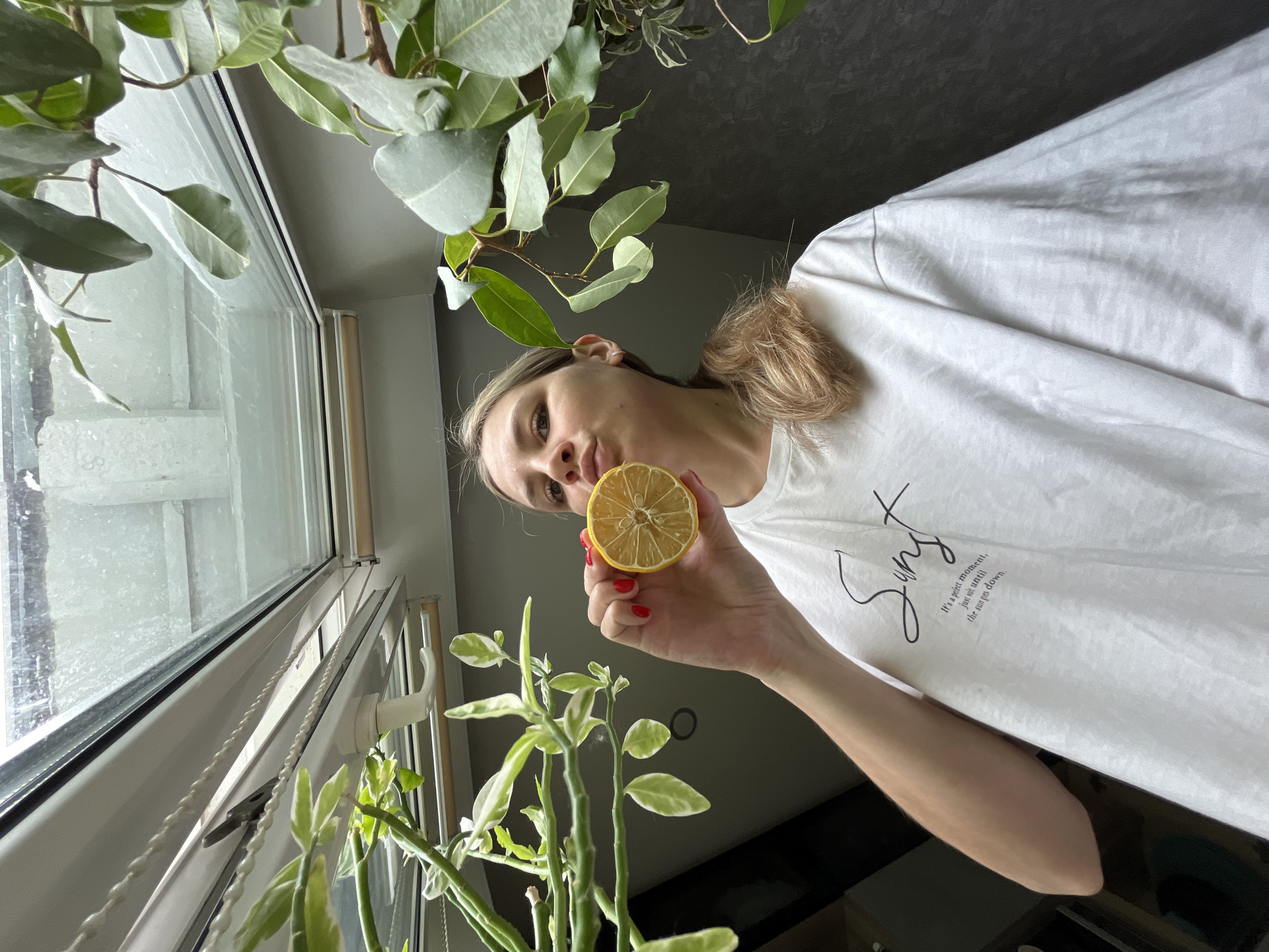 A young woman with blonde hair pulled back in a ponytail is holding a dried lemon slice up to her lips. She is wearing a white t-shirt with black text. The woman is standing in front of a dark gray wall, with a large window with a crank handle above her. Green plants are visible on the left and right sides of the frame, framing the scene. The image was taken indoors, likely in a residential setting. The provided image does not contain any specific information that would allow for its localization to Chaikovsky, Russia.