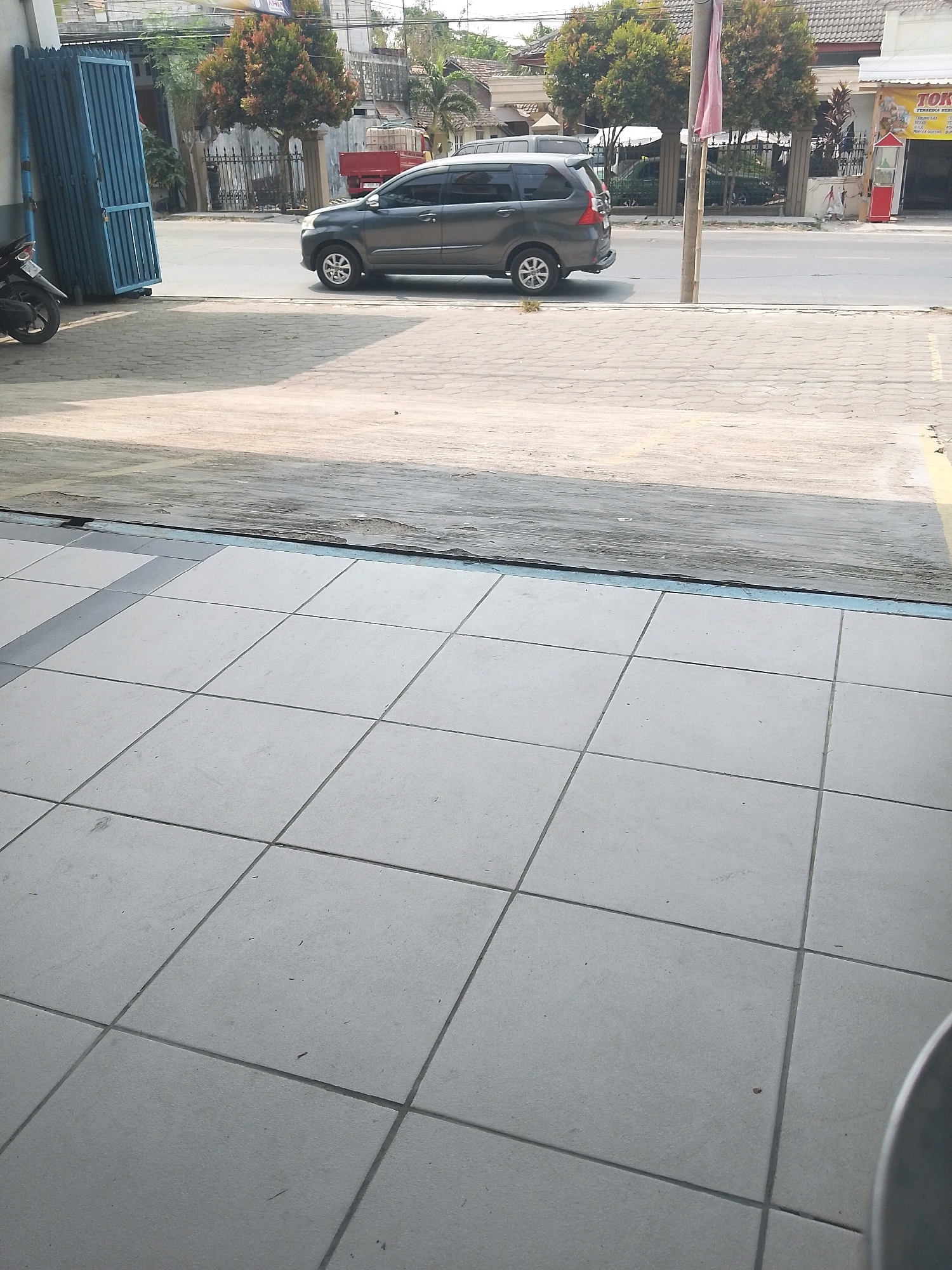 The image depicts a street scene in Kalitidu, Indonesia, during the daytime. In the foreground, a tiled walkway runs parallel to a paved road. On the road, a dark gray minivan is parked, facing left. Behind the minivan, a red truck is partially visible. To the left of the minivan, a blue folding gate stands closed, adjacent to a motorcycle parked at an angle. The background features buildings with pitched roofs, trees, and a street lamp. A white awning with yellow lettering, possibly indicating a shop, is visible on the right side of the frame. The overall atmosphere is one of a typical urban street environment.