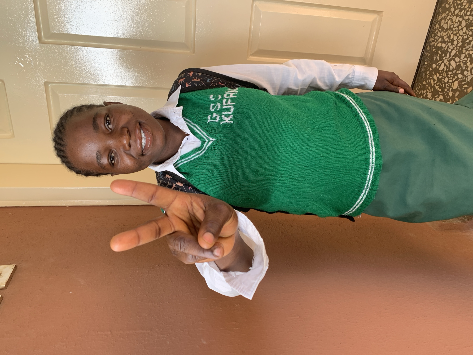A young woman smiles and makes a peace sign. She is wearing a green sweater vest with white trim and the words "GSC KUFAK" written on it. She is also wearing a white collared shirt. She is standing indoors with a light brown wall behind her and a white door to her left. It is possible the location is Jos, Nigeria based on the context of her sweater. The woman's hand is in the foreground making the peace sign.