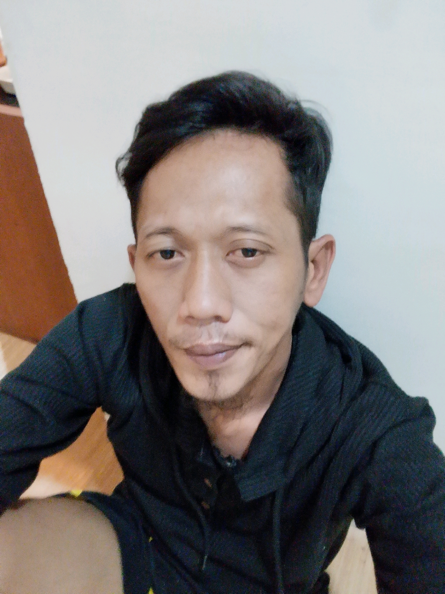The image is a close-up, selfie-style portrait of a man with short, dark hair and a light complexion. He is wearing a dark-colored hooded sweatshirt. The background consists of a plain, light-colored wall and a wooden floor with some reddish-brown tones. There is a visible shadow line on the left side of the image, suggesting the presence of an object or wall corner. The man is looking directly at the camera. The setting appears to be an indoor environment. No specific landmarks or contextual elements are visible to indicate a precise location within Tembilahan, Indonesia, beyond the general inference of an indoor residential or public space.