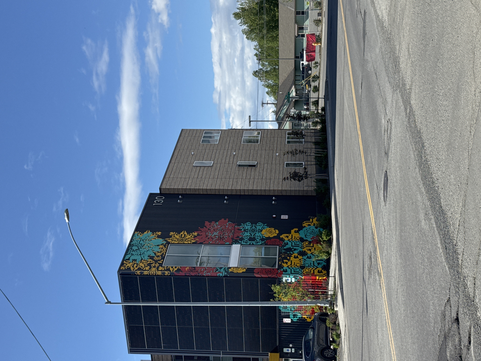 This is a street-level, eye-level, full shot of a modern building with a colorful mural on its side, located in Anchorage, Alaska. The building is dark grey with a section covered in wood-like siding. The mural features stylized floral patterns in yellow, teal, red, and black. The building has several windows, and the number "130" is visible on the dark grey section. A street lamp with a curved arm extends over the building. In front of the building, there is a dark grey car parked, and some landscaping with small trees and bushes. The foreground shows a paved road with yellow lane markings and some patches of wear. In the background, across the street, are several low-rise buildings, one of which is green, and some trees line the horizon under a blue sky with scattered white clouds. There are no people visible in the image.