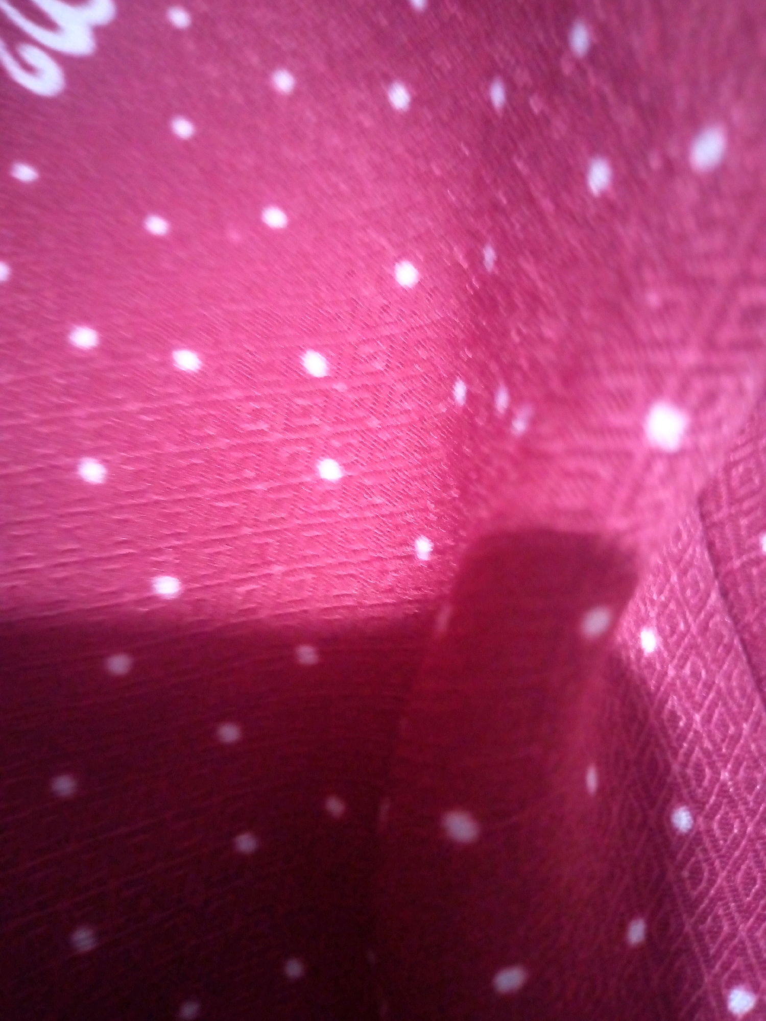A close-up shot shows a textured pink fabric with white polka dots. The fabric is folded and creased, creating shadows and highlights. The texture of the fabric appears to be woven, with a subtle diamond pattern visible in some areas. A portion of a white, cursive design is visible in the upper left corner of the image, partially obscured by the fabric. The lighting is uneven, suggesting the image was taken with a flash or under artificial light. There are no people or discernible environmental details present in the image.