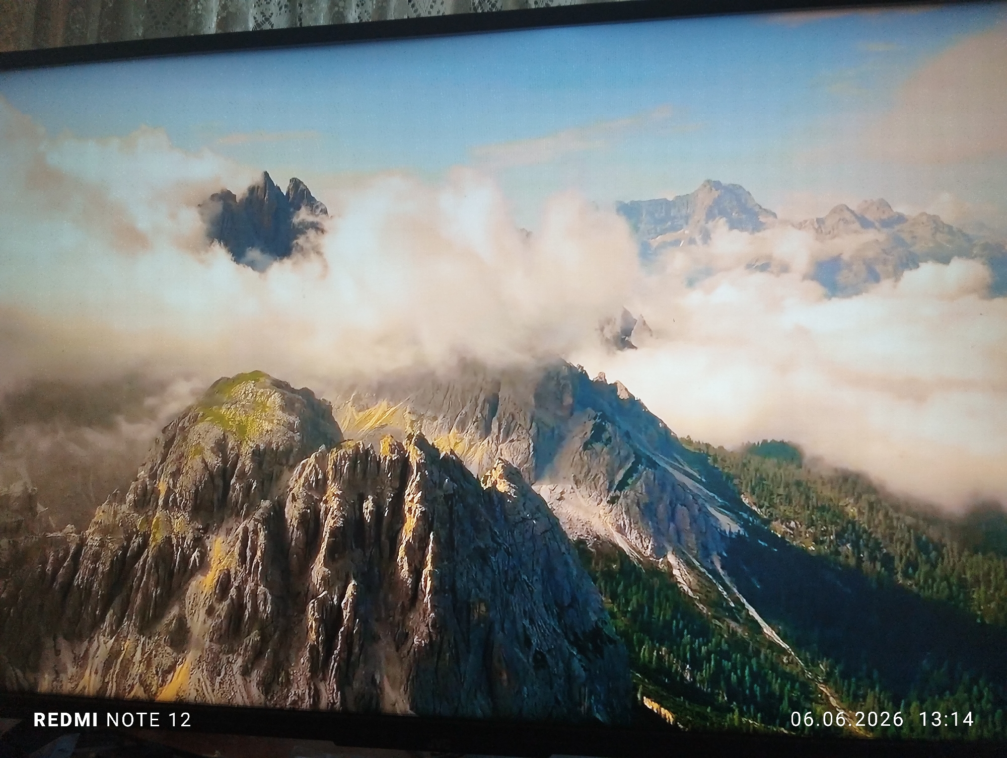 The image is a photograph of a screen displaying a panoramic mountain landscape, captured in the city of Burgas, Bulgaria. The depicted scene features a rugged mountain range characterized by rocky peaks and slopes, partially enveloped by extensive white and light brown clouds. The sky above is clear blue with scattered lighter clouds. Foreground and midground elements include prominent, steep rock formations exhibiting textures and patches of light green or yellowish vegetation, suggesting lichen or sparse plant growth. Coniferous forests densely cover the lower mountain slopes and valleys. Distant mountain peaks are visible through or above the cloud cover. The lighting casts distinct shadows and highlights, emphasizing the rugged topography. No people, animals, or artificial structures are present within the displayed landscape. Textual information is visible at the bottom of the screen: "REDMI NOTE 12" is positioned on the lower left, and "06.06.2026 13:14" is located on the lower right, indicating the device used to capture the image of the screen and the timestamp of that capture, respectively.