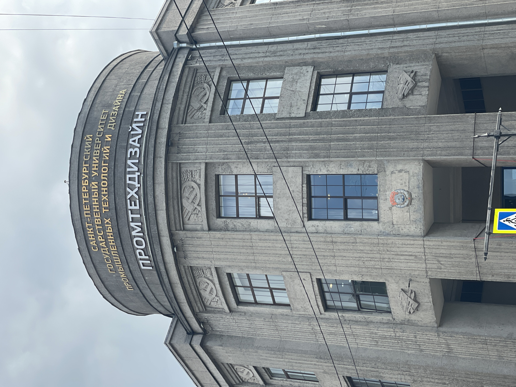 The image depicts the exterior of a building in Saint Petersburg, Russia. Specifically, it shows a section of the Saint Petersburg State University of Industrial Technologies and Design ("ПРОМТЕХДИЗАЙН" in Russian). The building's architecture is characterized by its gray, concrete facade with prominent horizontal ribbing. The curved section of the building is inscribed with the university's name in both gold and white lettering. Windows are visible, with black frames. The building also features stylized reliefs and ornamentation between the windows. Power lines bisect the frame, adding to the industrial context. A pedestrian crossing sign is present on the right side of the image. The sky is overcast.