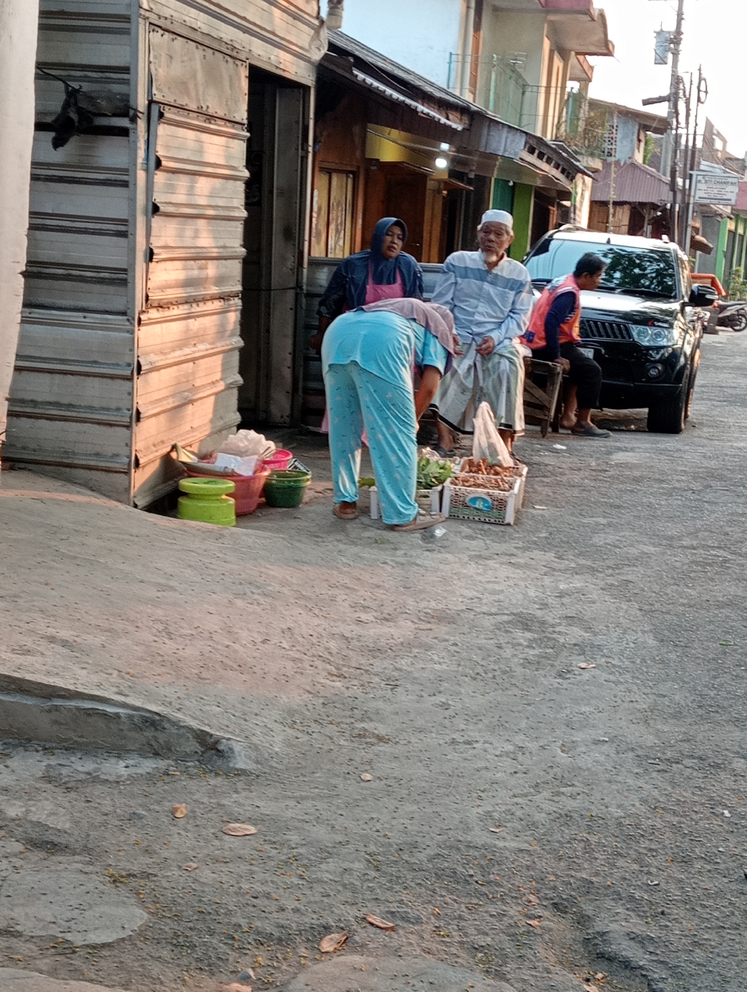 In Tanubayan, Indonesia, at street level, an outdoor scene unfolds near a weathered, metallic-sided structure. Three individuals are present. A person in a light blue top and matching pants is bent over, seemingly interacting with items on the ground. To their left, a person wearing a blue headscarf and a pink apron stands, looking towards the bent individual. To the right of the bent person, an elderly male in a striped long-sleeved shirt and white pants sits on a low wooden stool, facing forward. Behind him, another person in a reddish-orange vest sits on a similar stool, facing towards the right. A black SUV is parked on the right side of the frame. In the foreground, several plastic containers of varying sizes and colors (green, pink, and a smaller green one) are scattered on the ground. Two crates, one containing leafy vegetables and the other appearing to hold root vegetables, are also on the ground near the sitting individuals. The environment is a paved outdoor area, with buildings visible in the background. The lighting suggests it is either early morning or late afternoon, with warm sunlight casting shadows. Fallen leaves are visible on the ground.