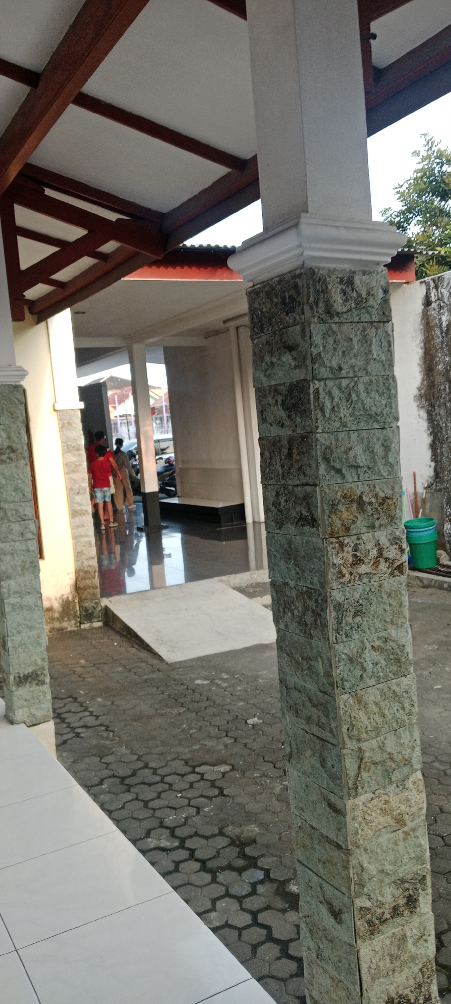 The image shows a covered walkway with a tiled floor and a stone-clad pillar in the foreground. In the background, several people are walking in a semi-enclosed space with a shiny, dark floor. There is a ramp leading up from the walkway into this space. The walkway has a wooden ceiling structure and white walls. To the right of the pillar, there is a concrete wall with some green buckets and other items. The overall impression is of an outdoor or semi-outdoor public area. The scene is located in the city of Kasihan, Indonesia.