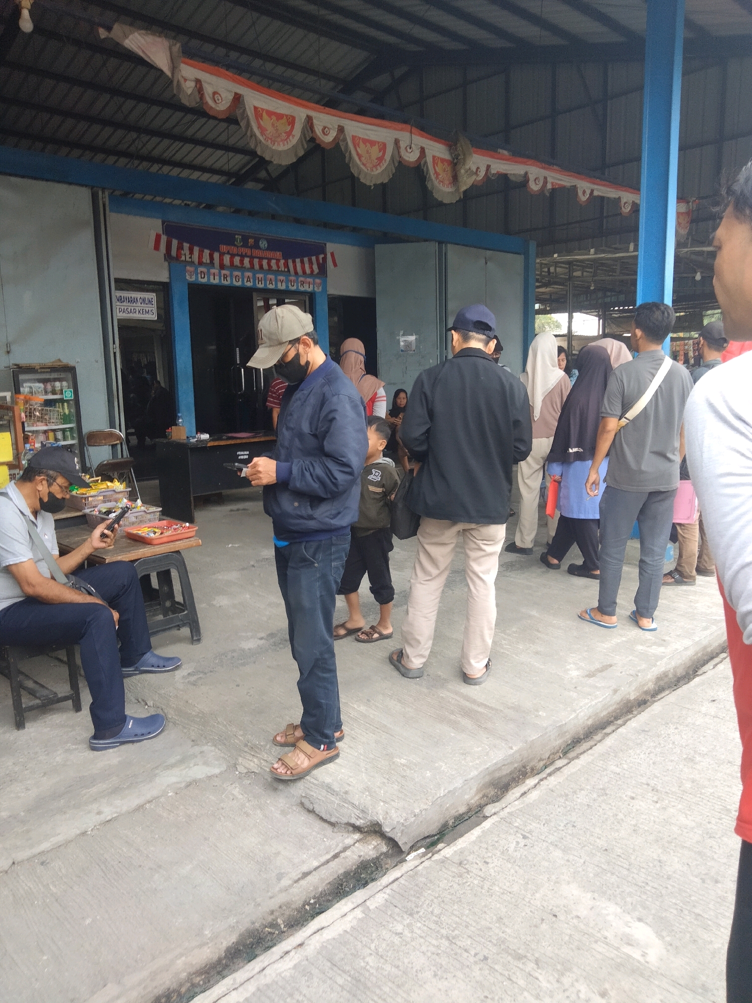 In Sukaasih, Indonesia, a group of people are gathered outside a building with a covered roof. The building appears to be a market or a government office, with signage that includes "PEMBAYARAN ONLINE" and "PASAR KEMIS". A banner above the entrance reads "DIRGAHAYU" with Indonesian flag motifs.
In the foreground, a man wearing a grey polo shirt and dark pants is seated on a chair, looking at his phone. Next to him, a man in a dark blue jacket and jeans is standing and also appears to be engaged with his phone. Several other individuals, including men, women, and children, are standing and walking in the background. Some are wearing traditional head coverings. The ground is paved concrete, and there are shelves with goods displayed near the entrance of the building. The overall scene depicts a public space with people engaged in various activities.