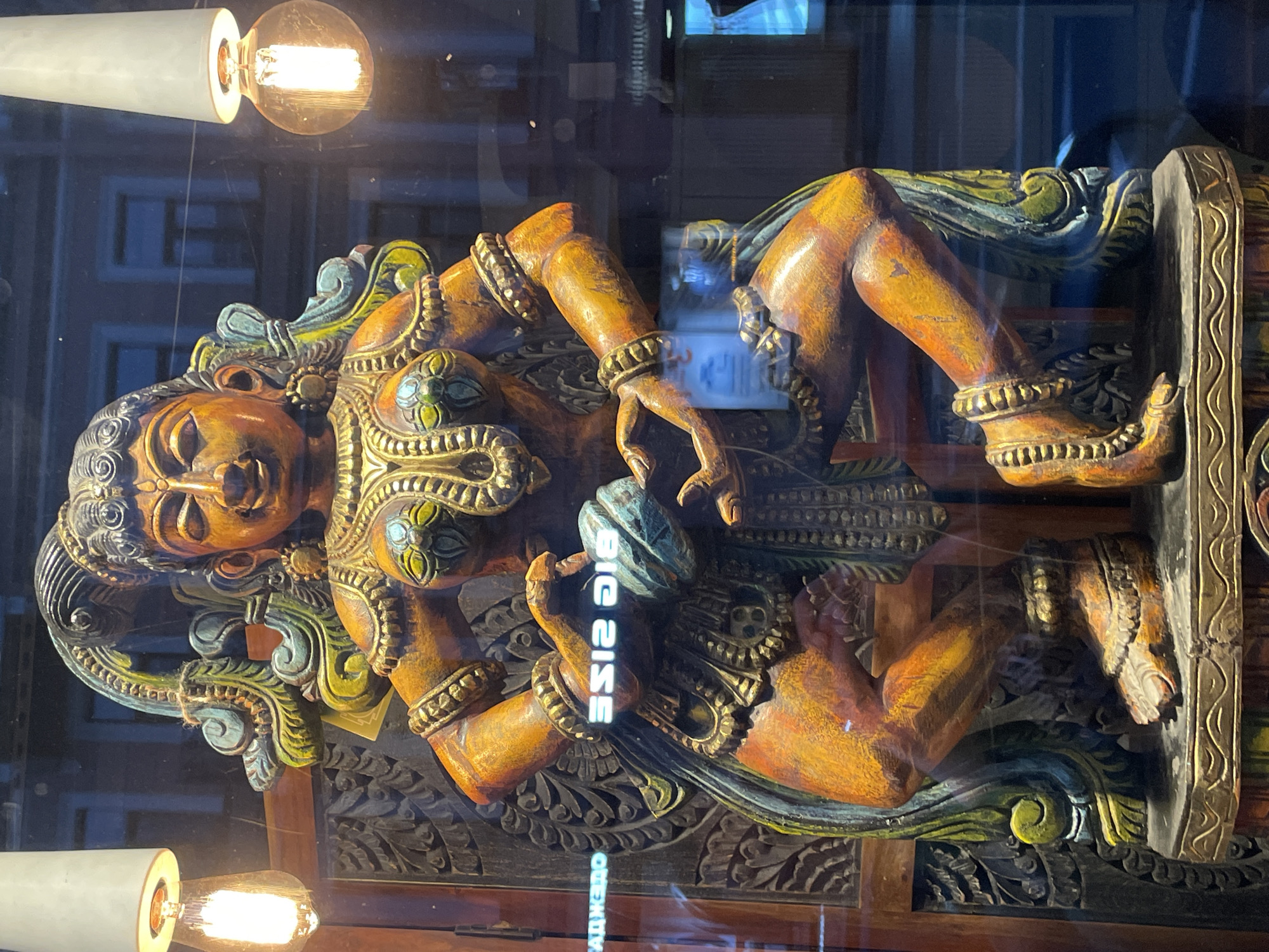 A color photograph shows a carved wooden figure in a display case, possibly in Saint Petersburg, Russia, as suggested by Cyrillic letters on a sign. The figure, painted in shades of orange, yellow, green, and gold, sits cross-legged with its head tilted upward. The figure has several bracelets on the arms and ankles. Decorative elements frame the figure, and the wood underneath is carved with intricate patterns.
The display case reflects an outdoor setting, with the tops of buildings visible. Two light fixtures with exposed bulbs are also visible at the top and bottom of the frame. A sign with the English words "BIG SIZE" is reflected on the glass, in reverse, along with text in Cyrillic.