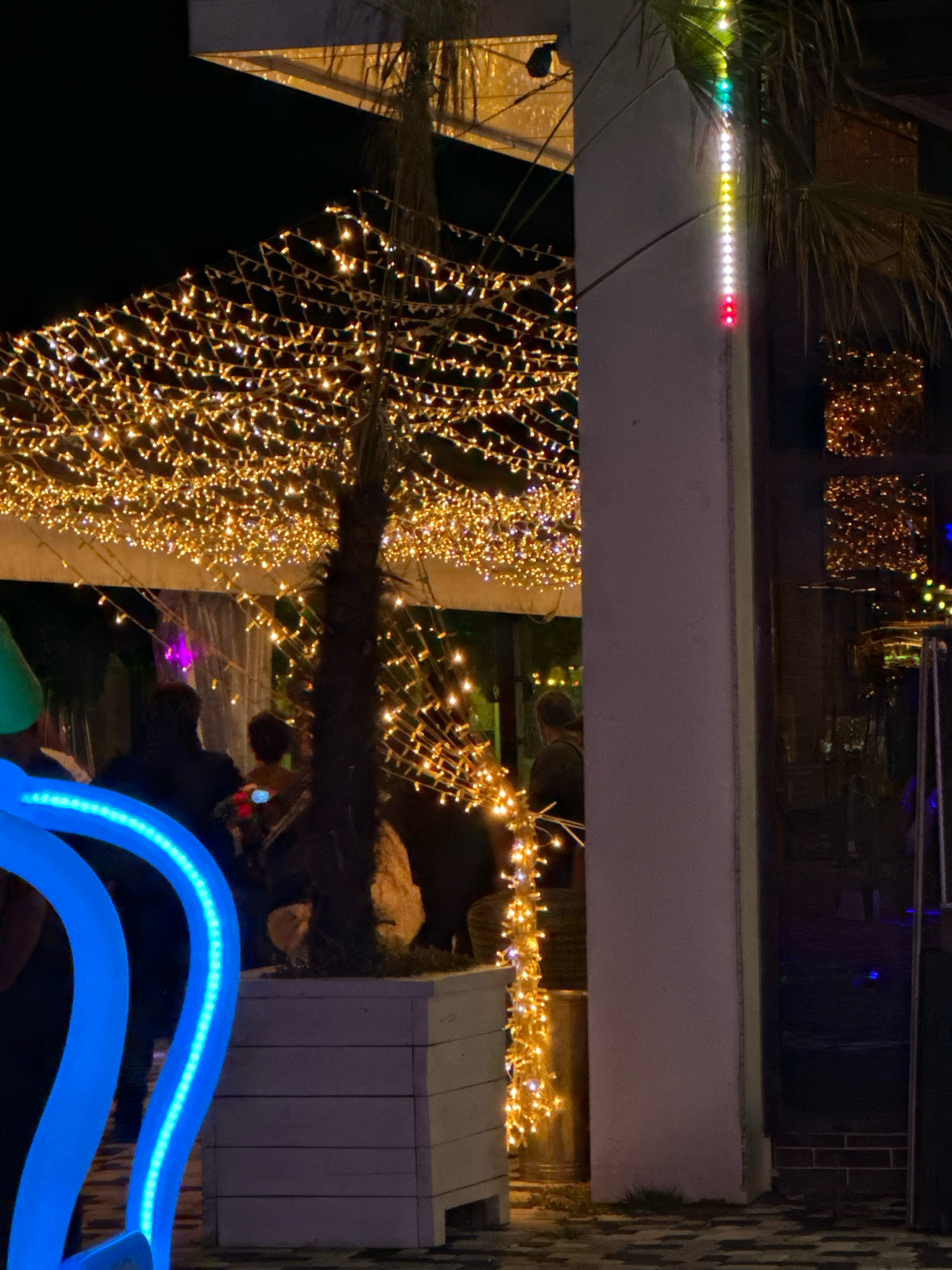 The image depicts a nighttime scene on a street in Lermontovo, Russia, likely a resort town. The foreground features a large, illuminated blue structure, possibly an art installation or decorative element, with a wavy, serpentine form. Behind this, a palm tree is visible, adorned with a cascade of warm-toned fairy lights that extend upwards and outwards, creating a canopy effect. To the right, a white pillar stands, illuminated by a vertical string of multi-colored LED lights, cycling through green, white, yellow, and red.
Several individuals are present in the mid-ground, mostly silhouetted against the bright lights. Some appear to be seated or standing in groups, suggesting a social gathering or dining area. One person on the far right is looking towards a reflective surface, likely a window or glass door. The ground is paved with dark cobblestones. The overall atmosphere is festive and illuminated, characteristic of an evening outdoor venue.