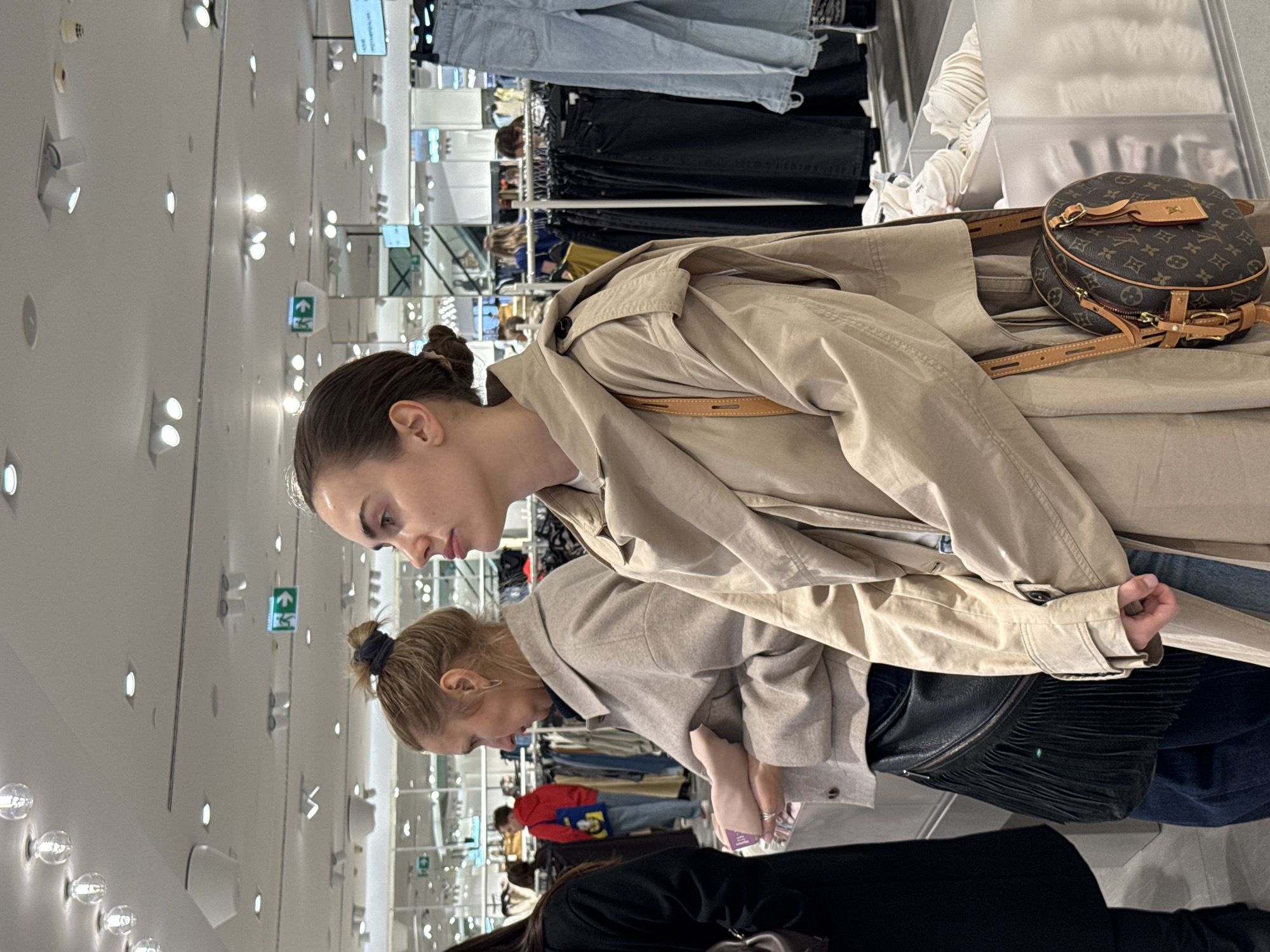The image depicts three individuals in what appears to be a clothing store in Unknown, Unknown. The primary focus is on two women in the foreground, both wearing light-colored coats. The first woman is wearing a beige trench coat with a brown Louis Vuitton bag slung over her shoulder. The second woman is wearing a light gray coat and a black fringe purse is visible on her shoulder. Below them, a third person wearing a black coat is partially visible. The environment appears to be a retail space with rows of clothing racks.
