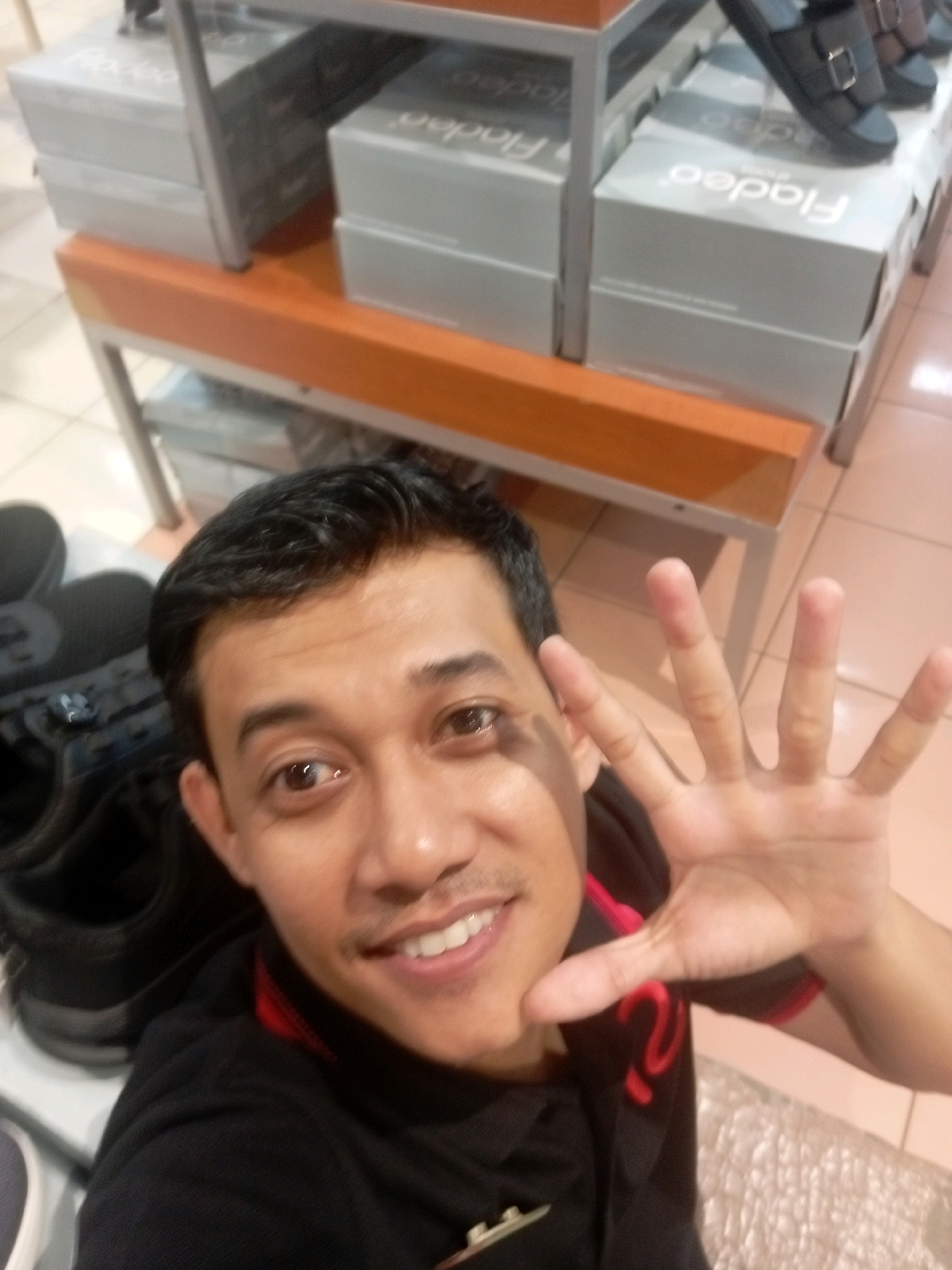 In Batam, Indonesia, a man with dark hair and brown eyes takes a selfie in what appears to be a shoe store. He is wearing a black shirt with red trim. His right hand is raised, fingers splayed, showing all five fingers. In the background, several shoe boxes are stacked on orange shelves. A pair of black sandals is visible on top of the boxes. The floor is tiled in a light brown color.