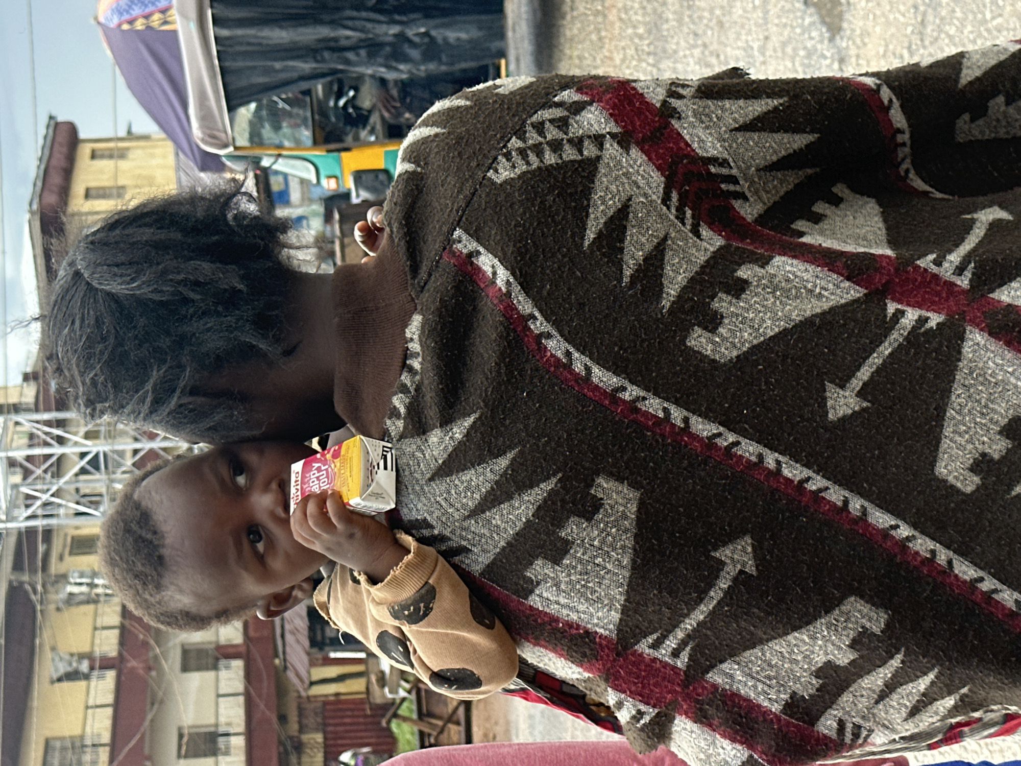 The image, taken in Gwong, Nigeria, shows a close-up of two people, likely a child and an adult, viewed from behind. The adult is wearing a patterned blanket or poncho with geometric designs in brown, gray, and maroon. The child is held close and is wearing a light brown sweatshirt with a dark polka dot pattern. The child is holding a small, yellow juice box with pink and white text. The text on the juice box is partially visible, with the words "happy hour" and some Korean characters discernible. The background suggests an outdoor, possibly urban setting with buildings and a vehicle resembling a tuk-tuk visible. The lighting indicates it's daytime. The framing of the shot, focusing on the subjects' backs and the child's interaction with the juice box, creates a sense of intimacy and everyday life.