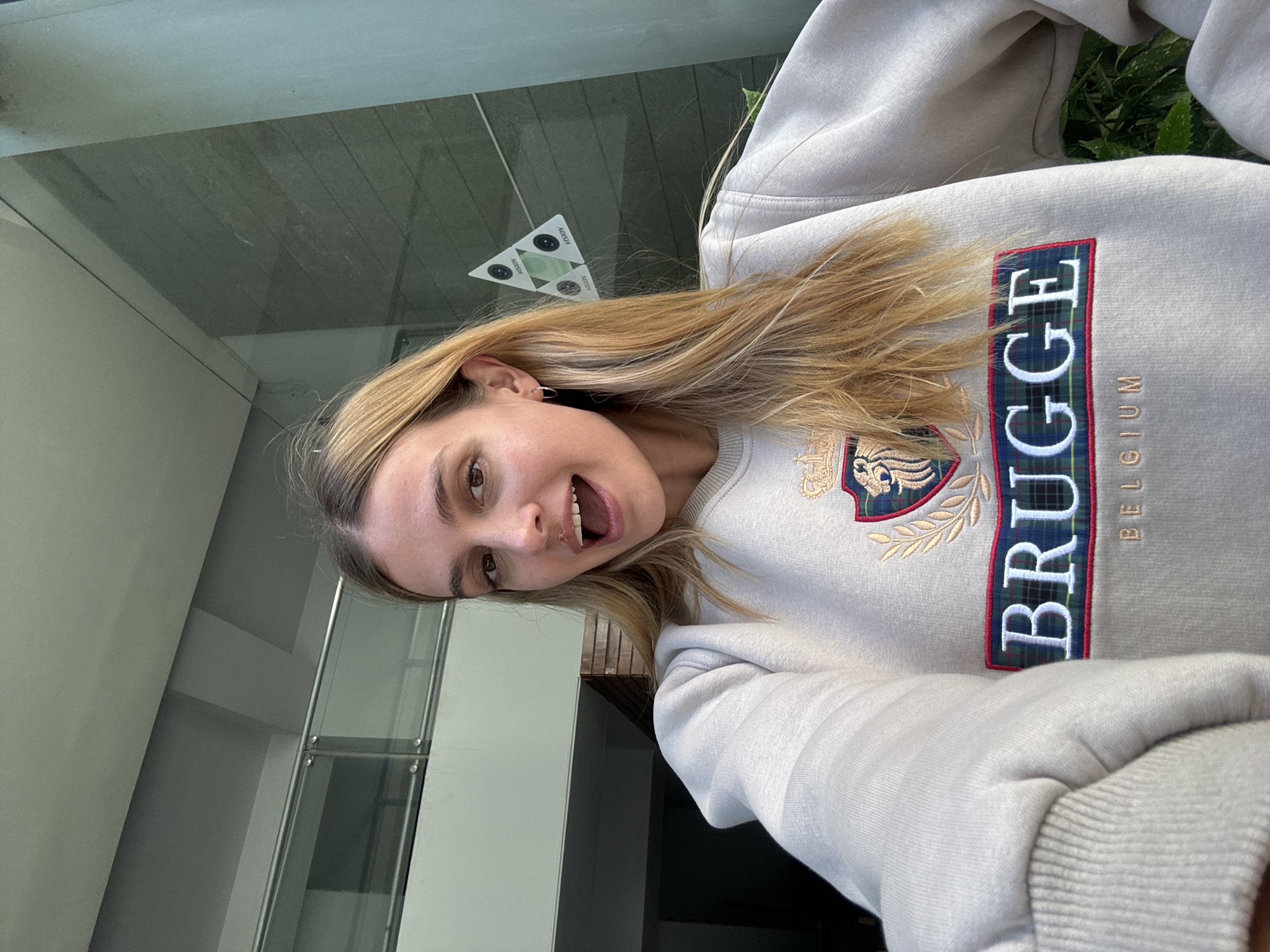 A young woman takes a selfie indoors. She is fair-skinned, and her hair is blonde. She wears a light-colored sweatshirt with the word "BRUGGE" vertically written in large, bold letters and the word "BELGIUM" beside it. She is smiling with her mouth open. To the left, architectural elements, potentially glass walls or a window frame, create an indoor environment. On the glass, there is a sticker with a triangular design. The location and city are not determinable from the image.