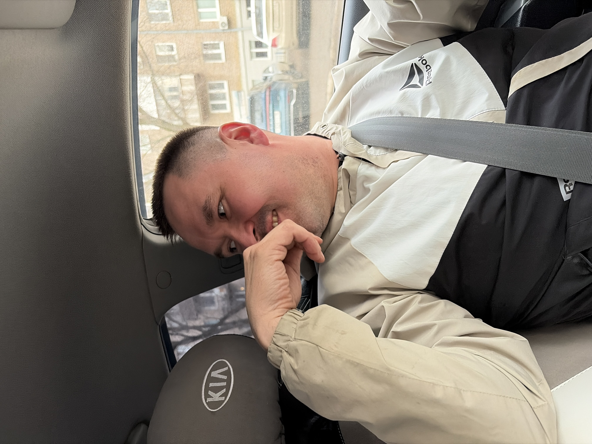 A man is seated in the back seat of a Kia car, wearing a Reebok jacket and a seatbelt. He is looking at the camera with a slight smirk, and his hand is near his face. Outside the car window, a building is visible. Based on the architectural style and signage, the location is likely Izhevsk, Russia.