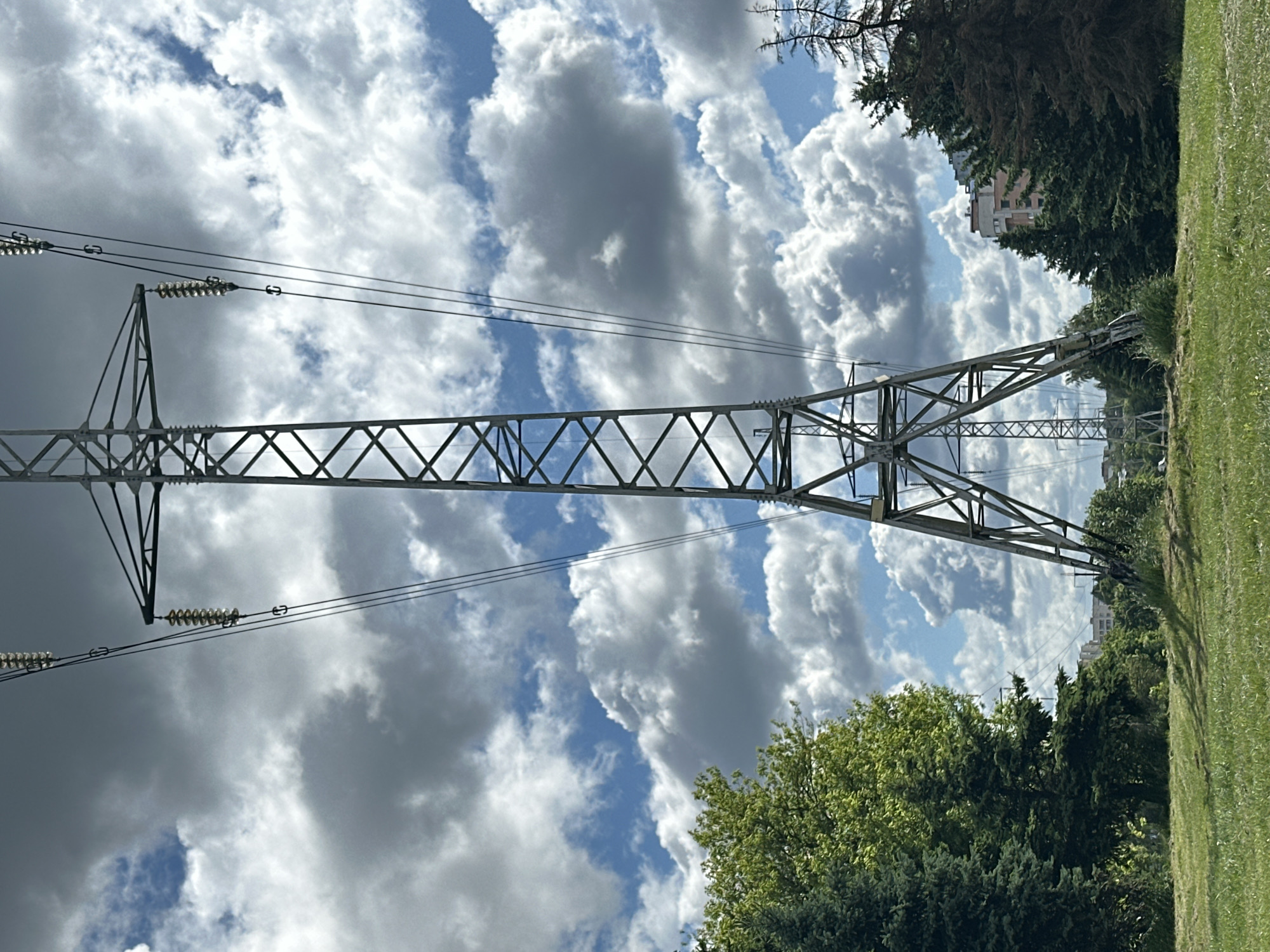 The image depicts a view looking upwards at a metal electricity pylon with transmission lines extending across a partly cloudy sky. The base of the pylon and its supporting structures are visible against a backdrop of trees and a grassy area on the right side of the frame. In the distance, a building is partially visible amongst the trees. The location is Minsk, Belarus.