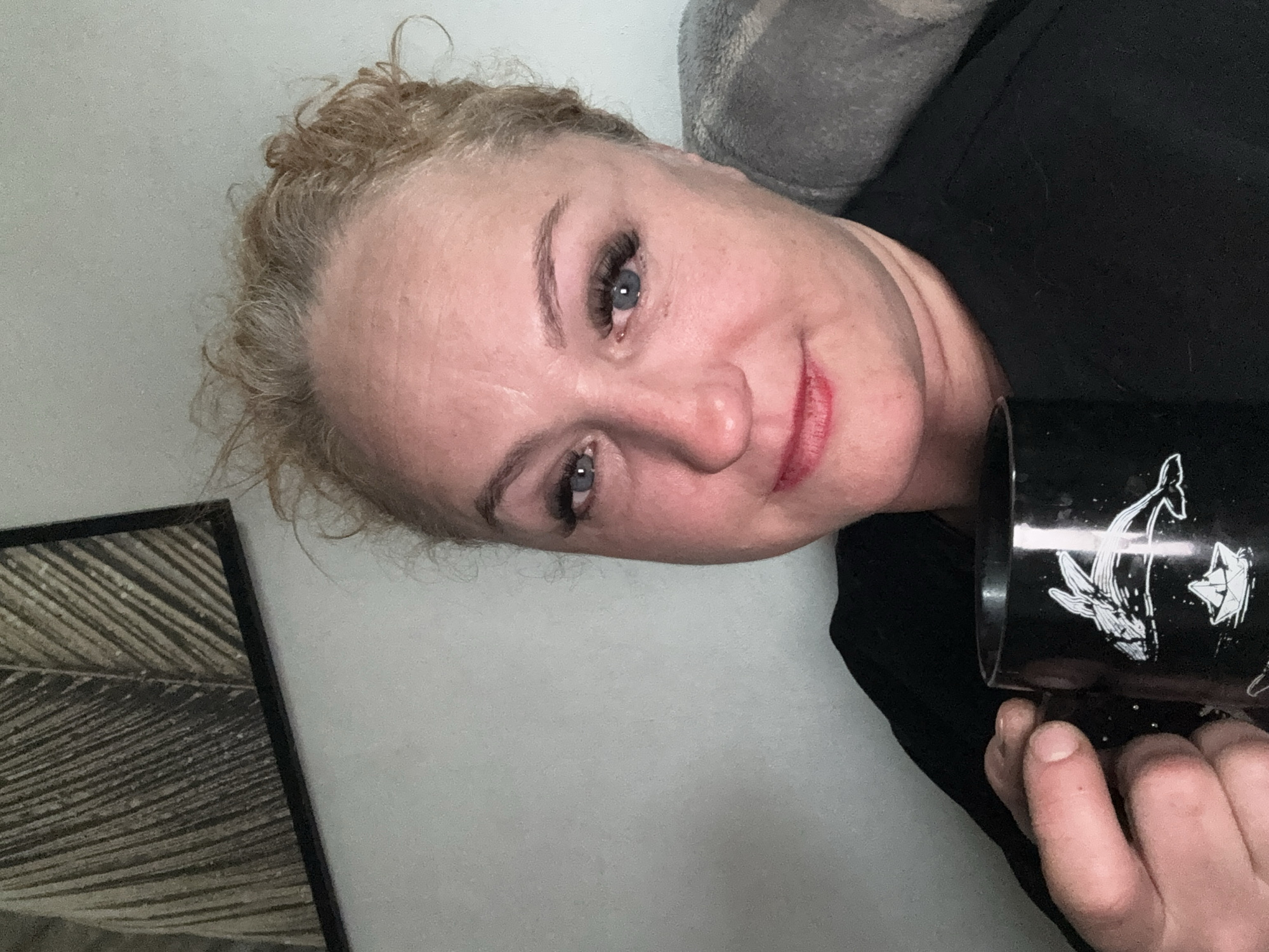A woman with blonde curly hair and blue eyes is lying on a light-colored surface, holding a black mug with white whale and origami designs. She is wearing a black shirt and has light makeup on her face, including lipstick and eyelash extensions. The background includes a grey pillow and a framed artwork with a textured pattern. The location is not discernible from the visual information provided.
