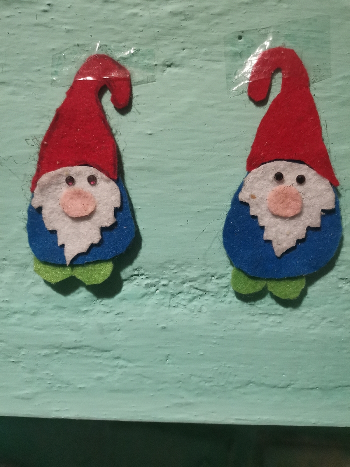 The image displays two handmade felt gnomes or dwarves affixed to a textured wall. The wall is painted in a light blue-green or mint green color and shows some surface irregularities and minor imperfections, suggesting an older or unevenly painted surface.
Both gnomes are nearly identical in design and coloring. Each gnome is crafted from layered felt pieces. They feature a prominent, elongated, and slightly curved red pointed hat. Below the hat is a white, fluffy-looking beard, covering the lower part of their face and extending down to their bodies. A small, light pink oval-shaped piece of felt forms the nose, centrally placed above the white beard. Two tiny, black circular beads or dots serve as their eyes, positioned above the nose and peeking out from under the hat, on the white beard piece.
The main body of each gnome is made from a rounded, blue piece of felt, visible below the white beard. Two smaller, rounded, lime green felt pieces protrude from the bottom of the blue body, representing their feet or shoes.
Each gnome is attached to the wall using pieces of clear adhesive tape, which are visible over the top, curved part of their red hats. The gnomes are positioned side-by-side, spaced apart, and appear to be facing directly towards the viewer. The lighting in the image is even, suggesting an indoor setting without strong directional light. No text is visible in the image.