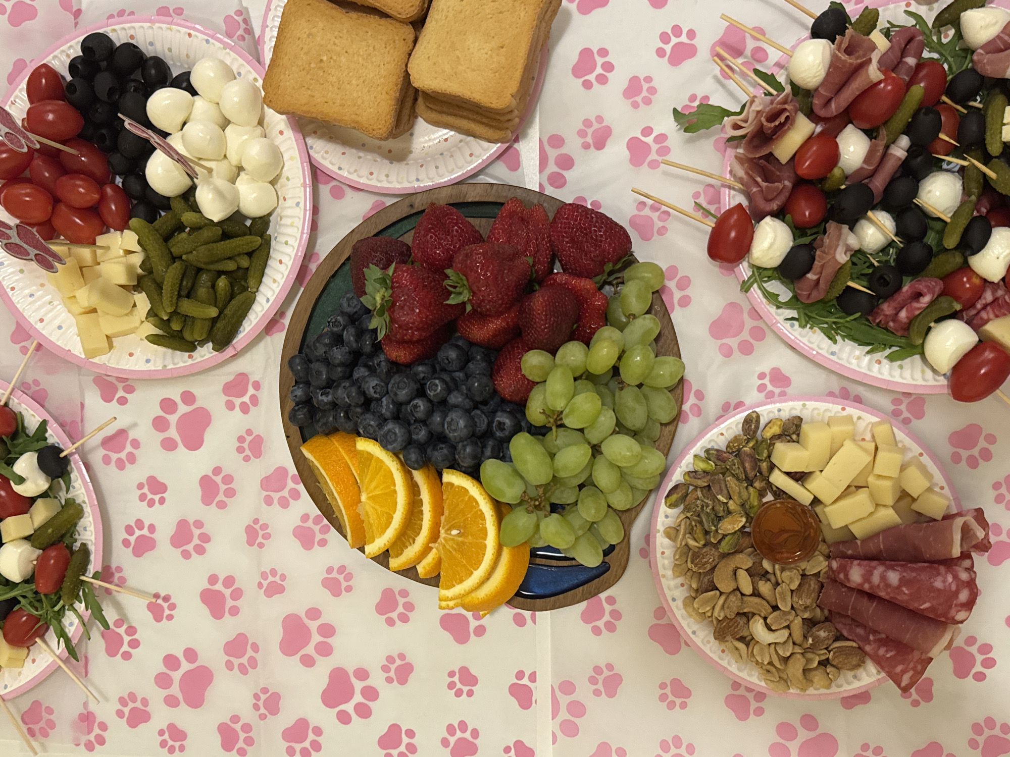 This overhead shot captures a vibrant spread of appetizers and snacks laid out on a white tablecloth adorned with numerous pink paw print patterns. The scene suggests a casual party or gathering, possibly with a pet or animal theme.
Several plates and a wooden board are filled with various food items. In the top left, a round paper plate holds distinct sections of red cherry tomatoes, shiny black olives, small white mozzarella balls (bocconcini), and a pile of small green gherkins (cornichons), alongside cubes of yellow cheese. Another paper plate directly above this one contains stacks of golden-brown rectangular toast or rusks.
The central feature is a large circular wooden board laden with fresh fruits: a generous amount of bright red strawberries, deep blue blueberries, bunches of green grapes, and slices of orange arranged along the edge.
To the right, another paper plate presents an assortment of individual skewers. These skewers are artfully arranged on a bed of arugula and feature combinations of cherry tomatoes, mozzarella balls, black olives, cornichons, and folded slices of cured meat (likely salami or prosciutto).
The plate in the bottom right corner offers a selection of dry snacks: a mound of shelled pistachios, a pile of mixed nuts (including cashews and almonds), cubed cheese, and folded slices of cured meat. A small round dish filled with an amber liquid, possibly honey or a similar condiment, sits among the nuts.
The overall impression is one of abundance and careful presentation, designed for easy snacking at a social event. No people or specific location cues are visible, and there is no discernible text in the image.
