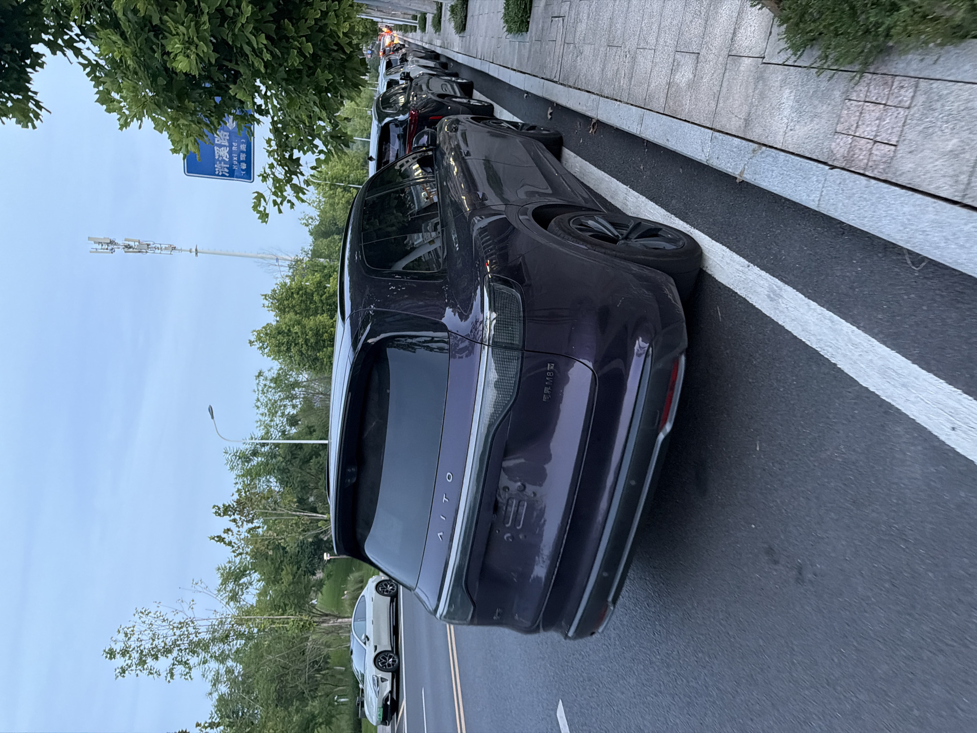 An AITO M7 electric SUV, colored dark purple, is parked along a street in Jiangjin, China. The vehicle is positioned in the foreground, with its rear facing the viewer. To its left, a white electric sedan is visible on the road. A row of parked vehicles extends down the street on the right side of the image. A blue street sign with Chinese characters and "Huxi Rd." is mounted on a pole to the right. A cell tower is visible in the background. The environment appears to be an urban or suburban area with trees and a clear sky.