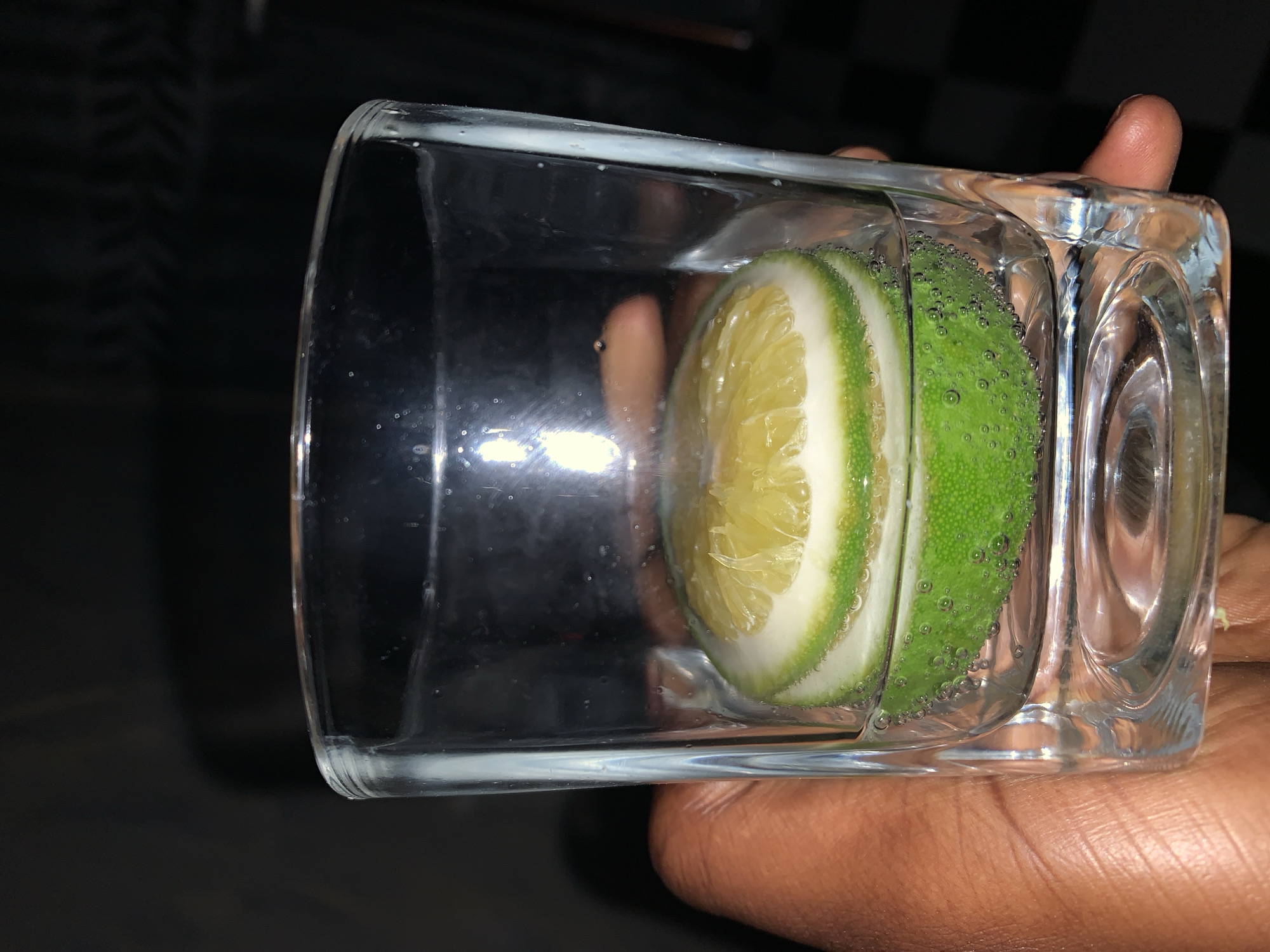 A hand holds a rectangular glass filled with a clear liquid, likely water. Inside the glass is a slice of lime, positioned so that the cut side faces the viewer. Small bubbles cling to the lime's rind. The scene appears to be indoors, with a dark background. It is not possible to verify the location as Rafin Par, Nigeria.