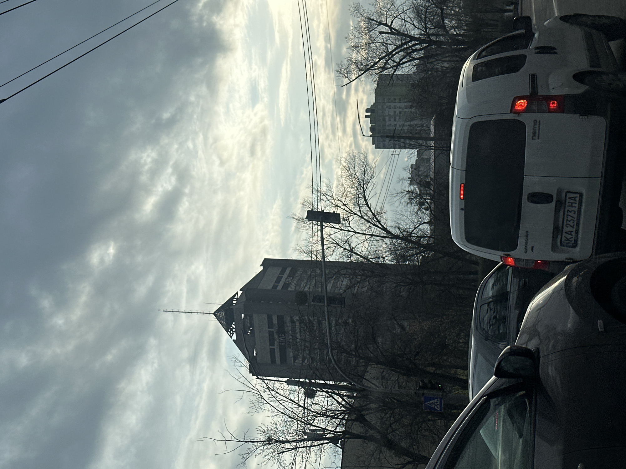 The image shows a street scene in Kyiv, Ukraine. A large, gray building with a spire is visible in the background. The sky is overcast with white and gray clouds. Utility poles and bare trees are in the foreground. Several vehicles are visible in the right half of the image, including a white van with the license plate "KA 2373 HA" and a dark-colored car. The overall impression is of a typical urban environment on a cloudy day.