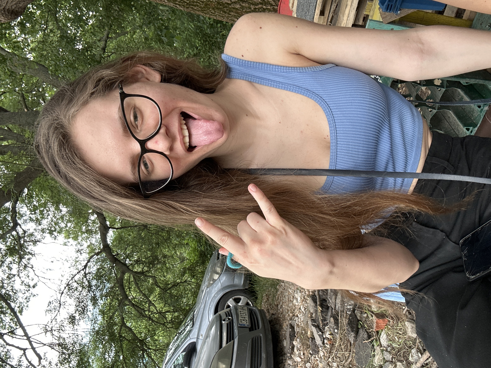 A young woman with long light brown hair and black-framed glasses is depicted outdoors, posing for the camera. She is smiling broadly, sticking her tongue out, and making a "rock on" hand gesture with her left hand, which has a light blue ring on the ring finger. She wears a light blue ribbed tank top and black pants. A smartphone with a cracked screen rests on her lap. The background features dense green foliage and tree trunks, indicative of a natural, wooded environment. To the left, two grey cars are partially visible; the closer vehicle bears a Bulgarian license plate "CB 4137 AH," confirming the location in Sofia, Bulgaria. The ground is composed of dirt, gravel, and broken asphalt. Stacked wooden pallets and a blue plastic crate are partially visible on the far right.