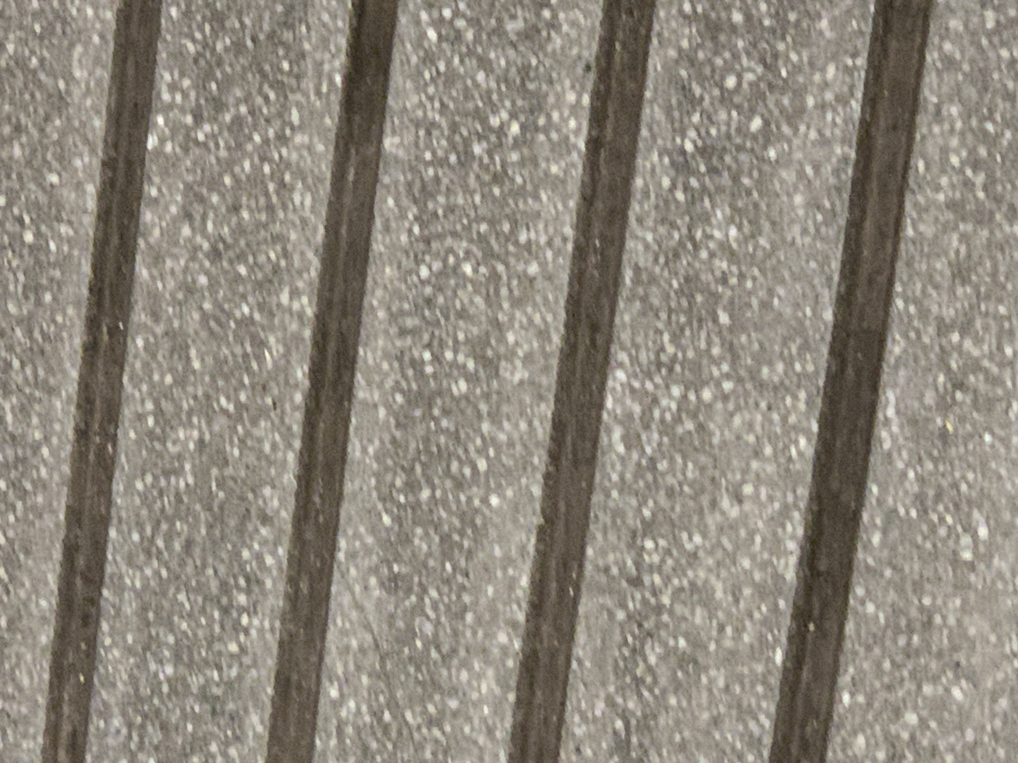 The image shows a close-up of stone flooring with recessed dark lines running diagonally across the frame. The stone appears speckled with light and dark gray tones. The scene appears to be outdoors. There is no evidence of people, subjects, actions, interactions, or specific objects beyond the stone flooring and recessed lines. The location is undetermined.