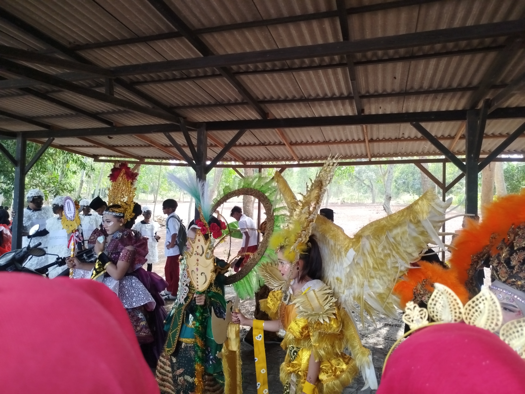 The image depicts a parade or cultural event in Jambu, Indonesia, taking place outdoors under a corrugated metal roof supported by wooden beams. Several individuals are dressed in elaborate costumes. In the foreground, a person in a bright red garment partially obscures the view. To their right, a person wears a golden, feathered wing costume with a yellow sash. To the left of this figure, another individual is adorned in a green and gold costume with a decorative mask and a tall headdress. Further back, more people in white, fluffy costumes and traditional attire are visible. In the background, trees and natural foliage suggest a rural or semi-rural setting. A motorcycle is parked on the left side of the image. One person in the background appears to be observing the proceedings.