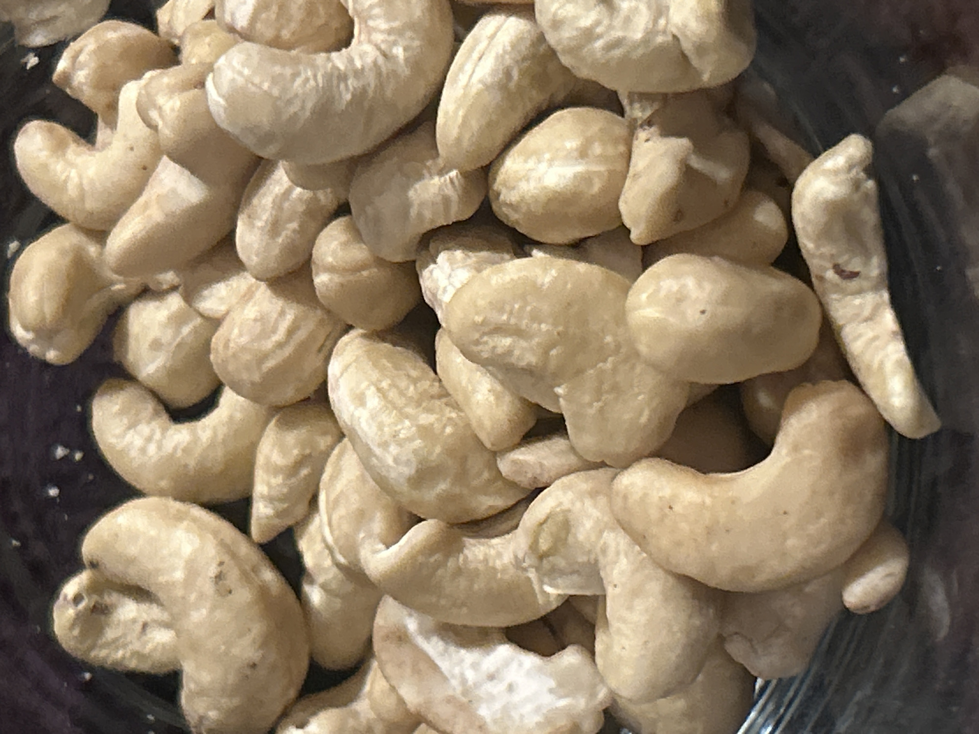 This is a close-up image of a pile of cashews. They are tan in color and appear to be unroasted. The cashews are tightly packed together. The scene is indoors, possibly a kitchen or pantry, but the specific location is unknown.