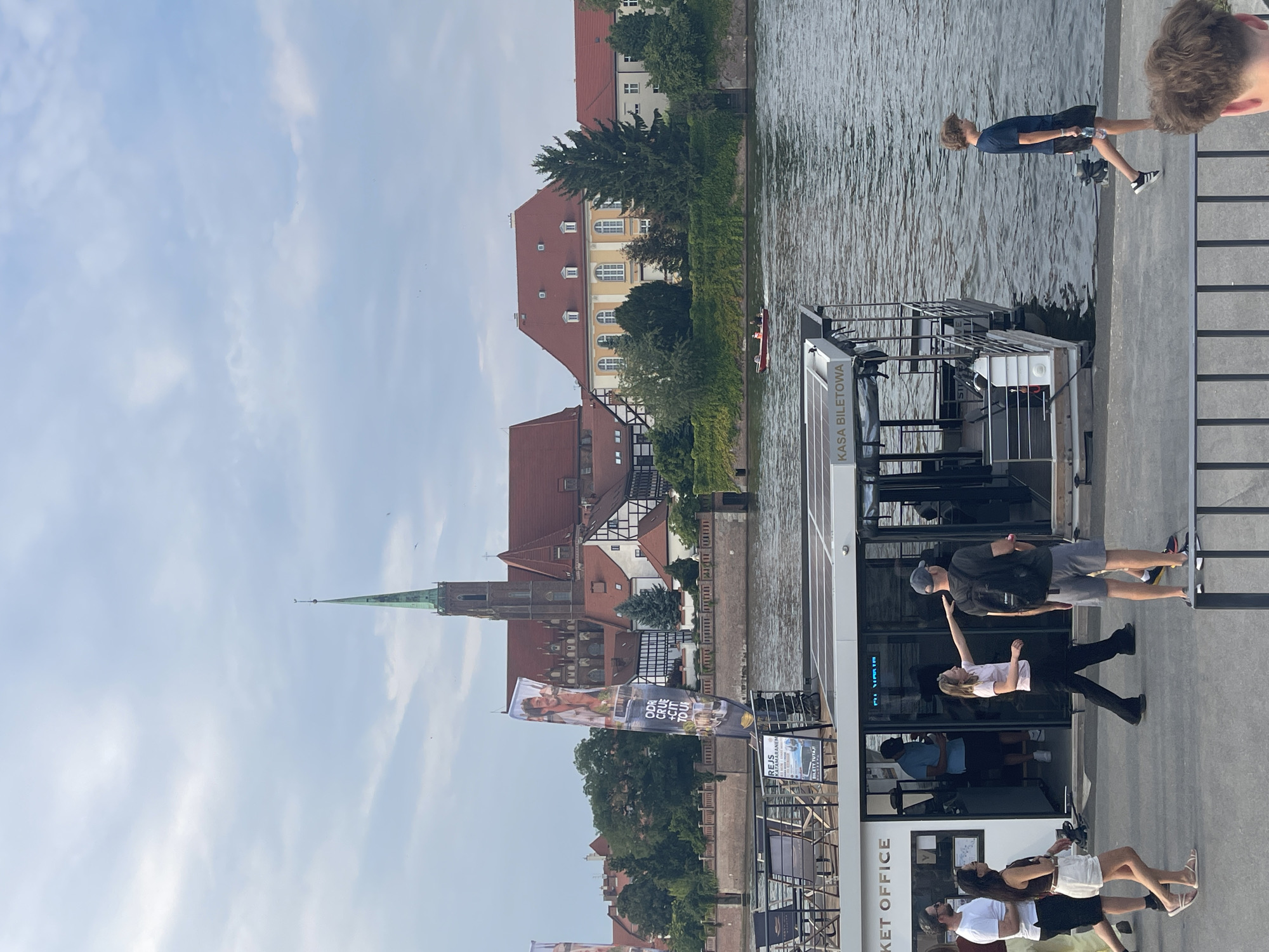The image captures a scene along the Oder River in Wrocław, Poland, on a cloudy day. In the background, a prominent green-spired cathedral dominates the skyline, with a complex of red-roofed buildings characteristic of historic European architecture situated behind a stone embankment. Trees and lush greenery line the riverbank. In the foreground, a modern pontoon boat with a "KASA BILETOWA" (Ticket Office) sign is docked. Several people are milling about on the walkway beside the boat. On the left, a man in a white t-shirt and shorts walks away from the camera, while a woman in a dark top and white shorts is looking back. Another woman in a pink top and black pants gestures towards the boat. A man with a backpack and shorts stands facing away from the camera. Further to the right, a young boy in a dark t-shirt and shorts walks along the waterfront, holding a water bottle. The river itself appears calm, with a small red boat visible in the distance.